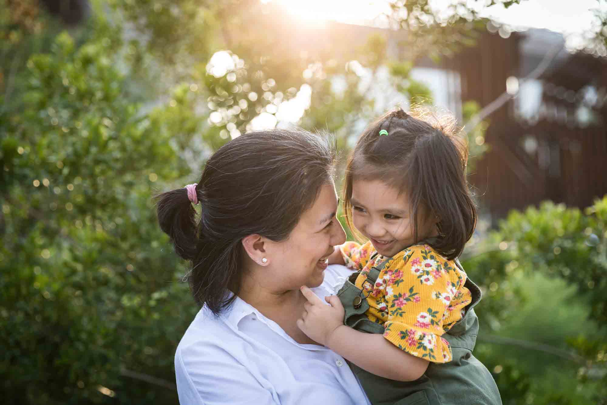 Mother holding young daughter in front of trees and bridge for an article on Brooklyn Bridge Park family portrait tips for young children