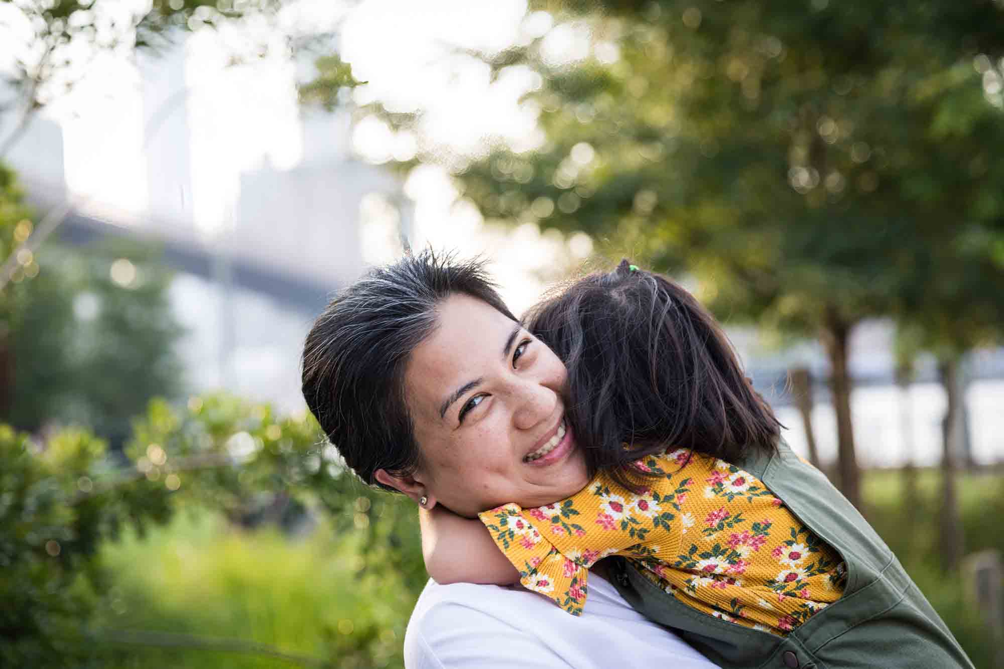 Mother holding young daughter in front of trees and bridge for an article on Brooklyn Bridge Park family portrait tips for young children