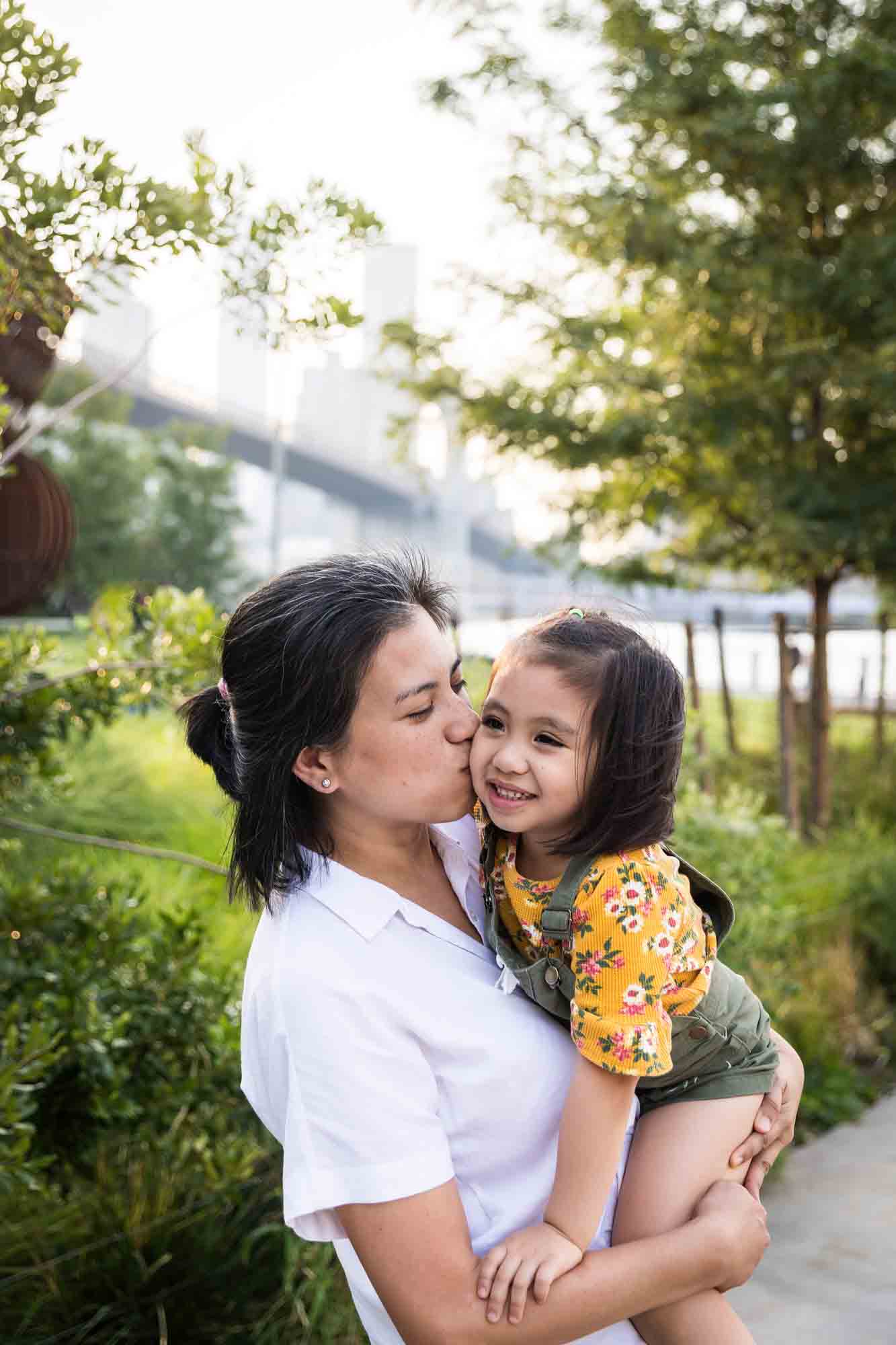 Mother holding young daughter in front of trees and bridge for an article on Brooklyn Bridge Park family portrait tips for young children