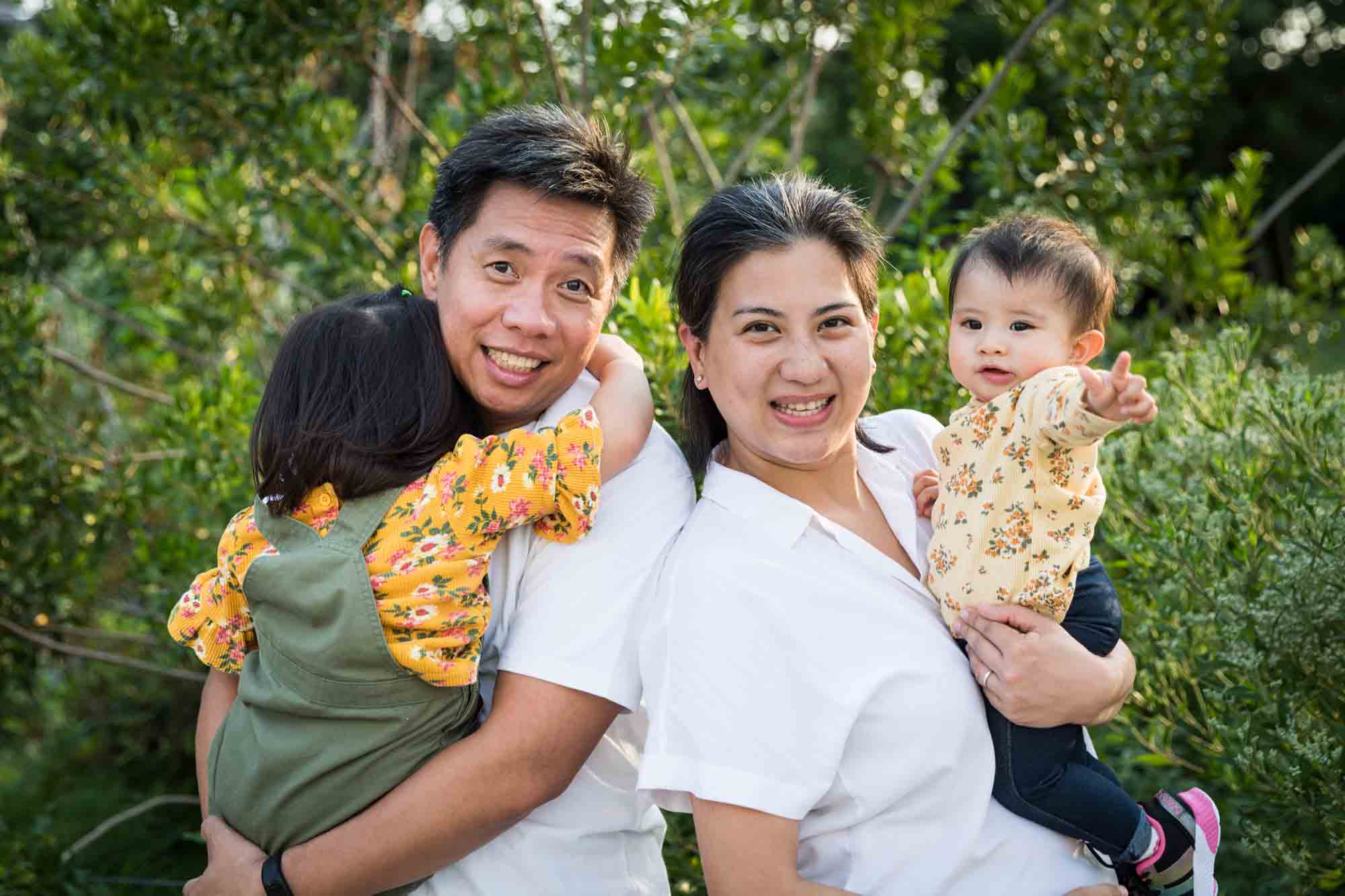 Parents holding young daughters in front of trees for an article on Brooklyn Bridge Park family portrait tips for young children