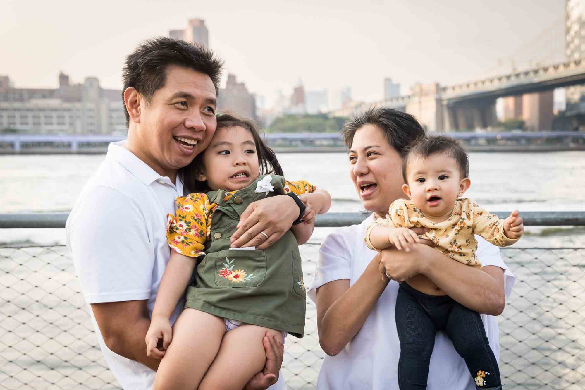 Parents holding two young children in the air in front of waterfront for an article on Brooklyn Bridge Park family portrait tips for young children