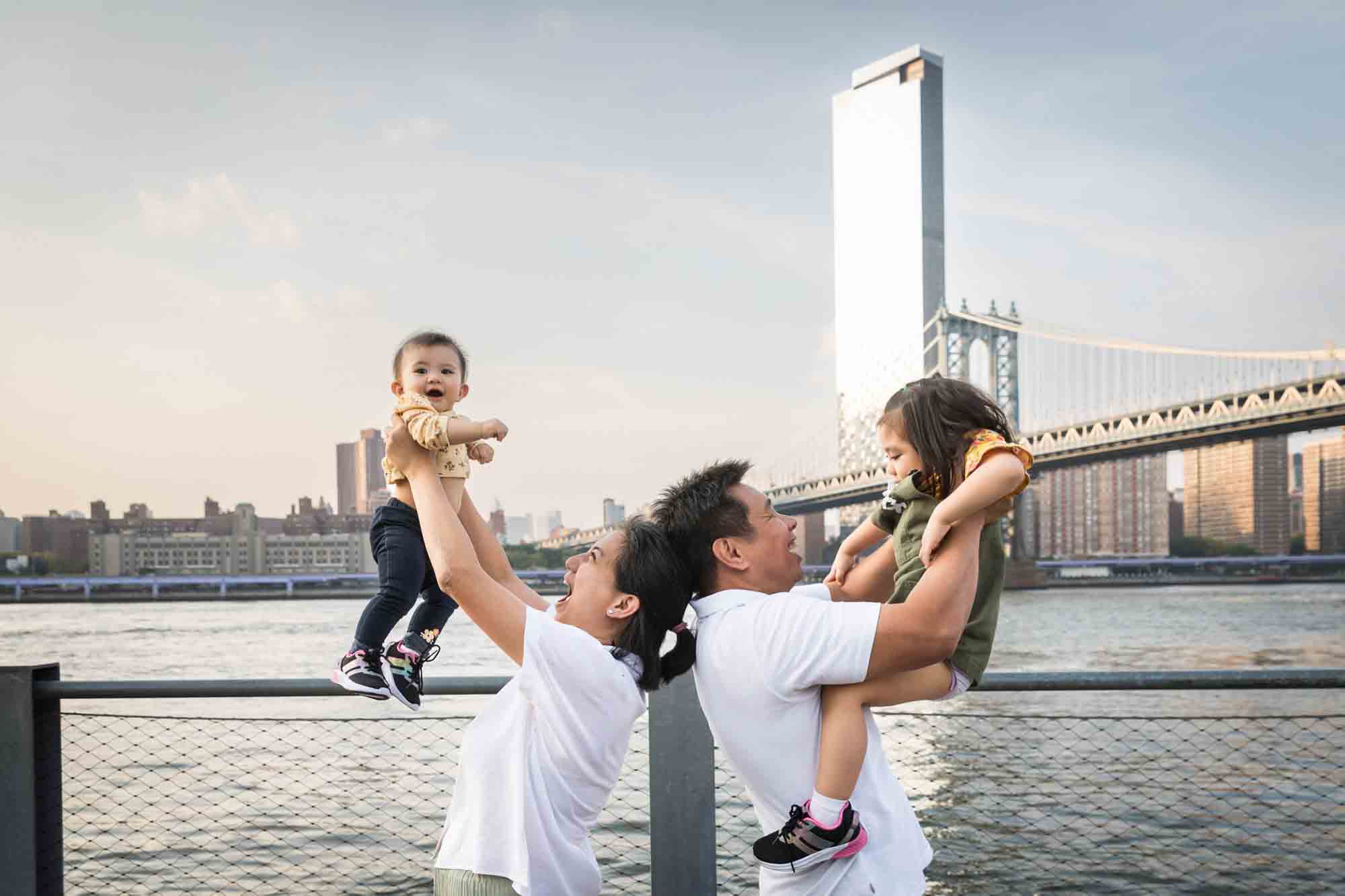 Parents holding two young children in the air in front of waterfront for an article on Brooklyn Bridge Park family portrait tips for young children