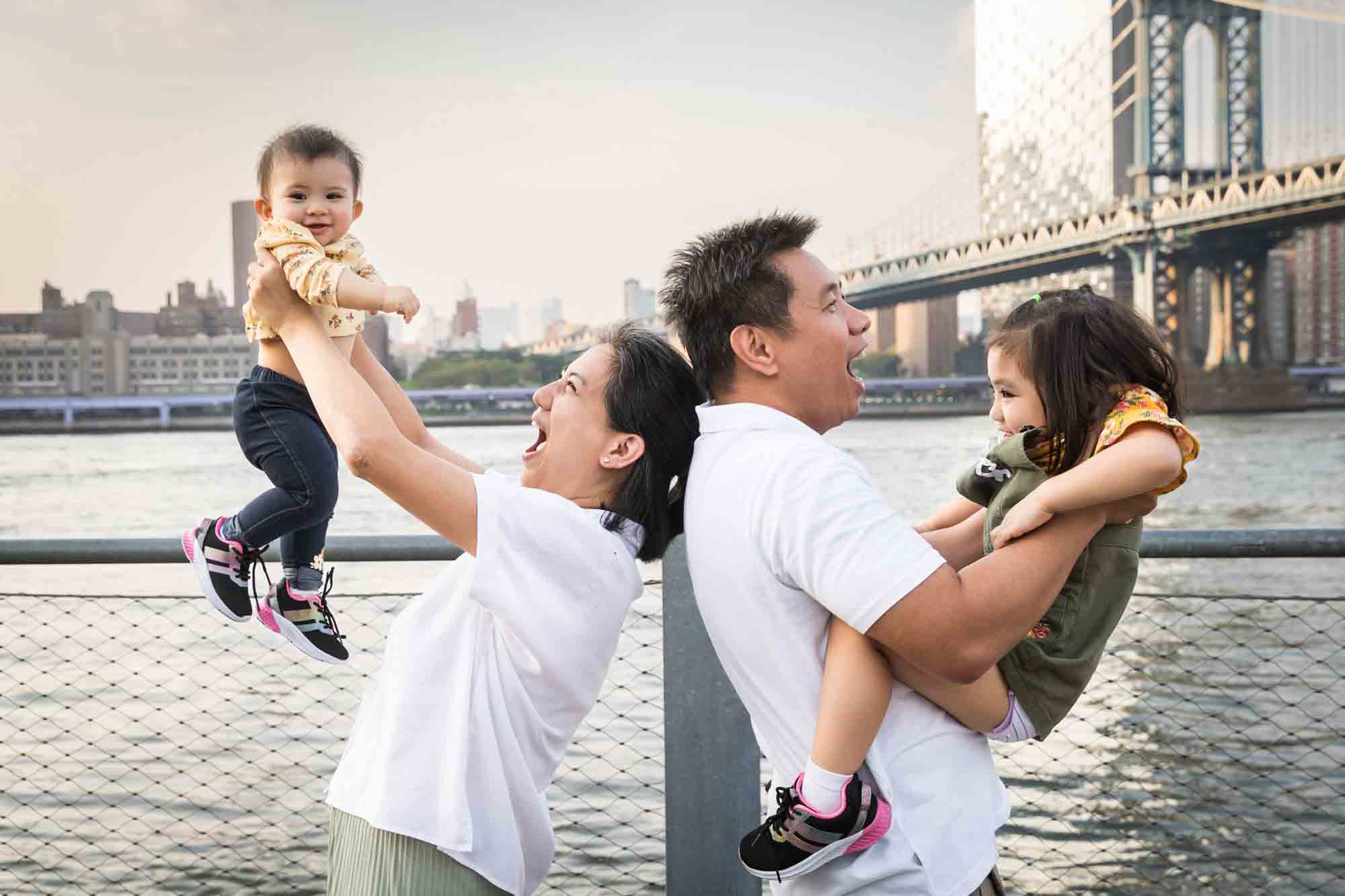 Parents holding two young children in the air in front of waterfront for an article on Brooklyn Bridge Park family portrait tips for young children