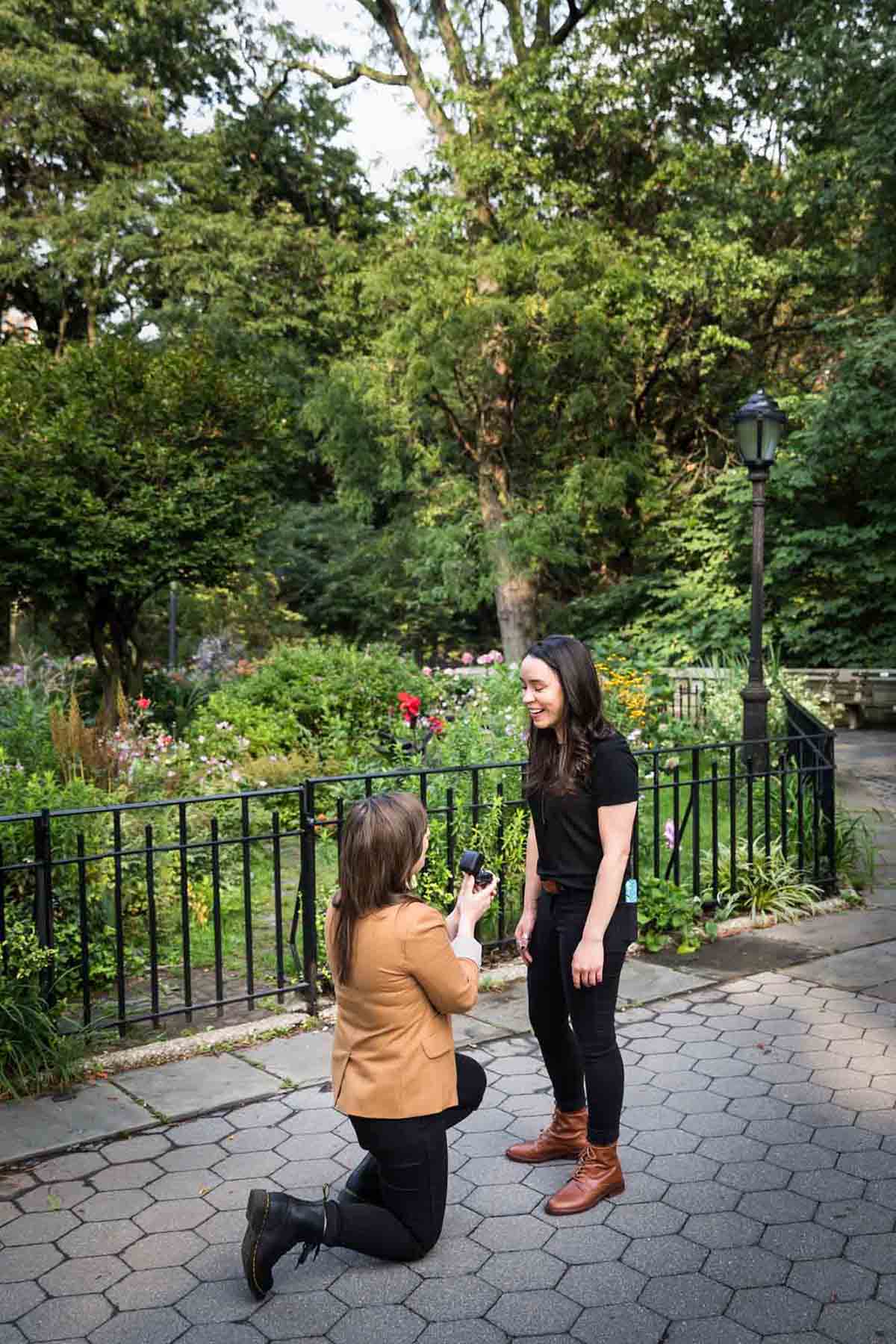 Woman on one knee showing engagement ring to another woman during Riverside Park surprise proposal