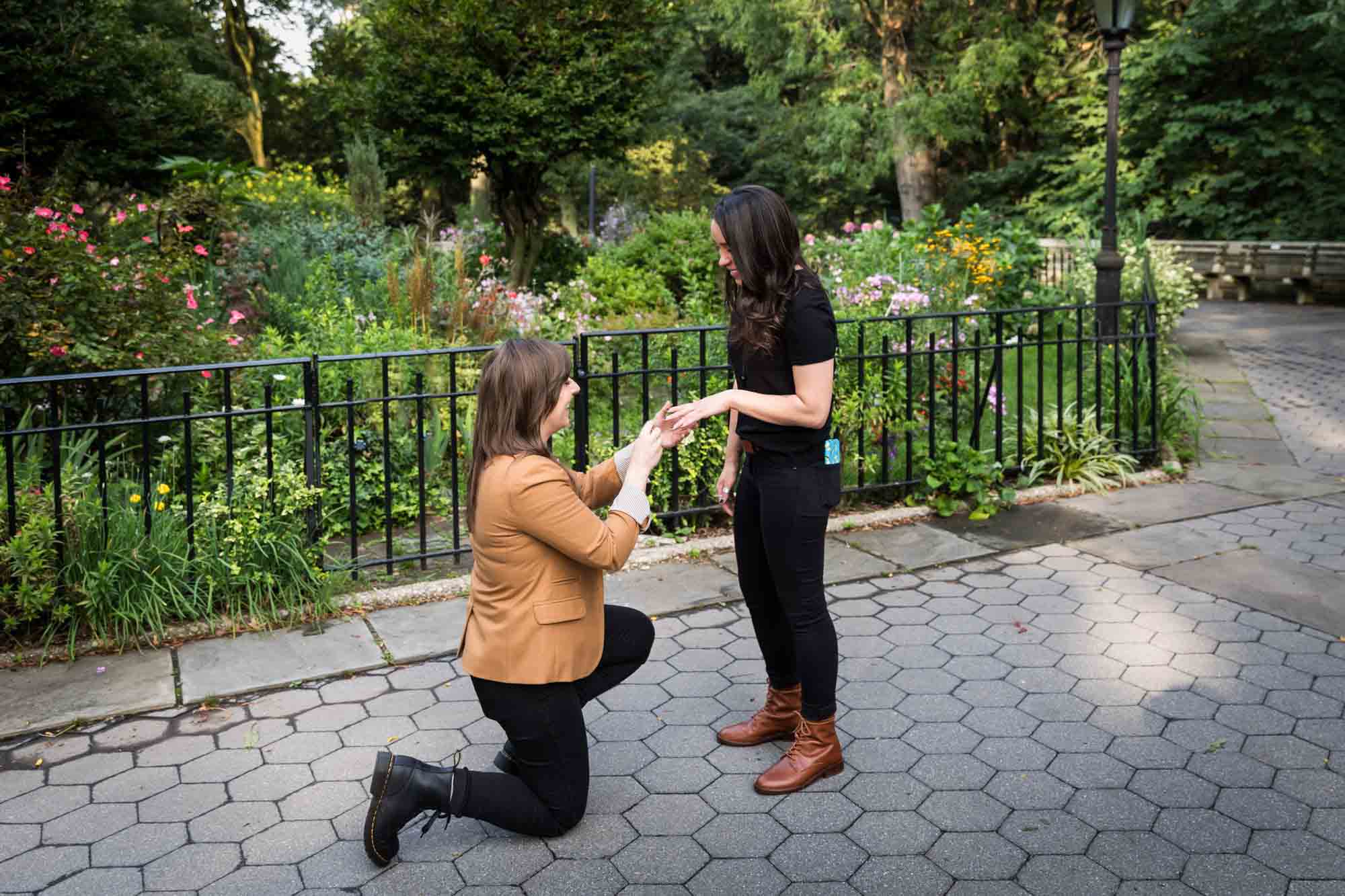 Woman on one knee putting engagement ring on another woman's hand during Riverside Park surprise proposal