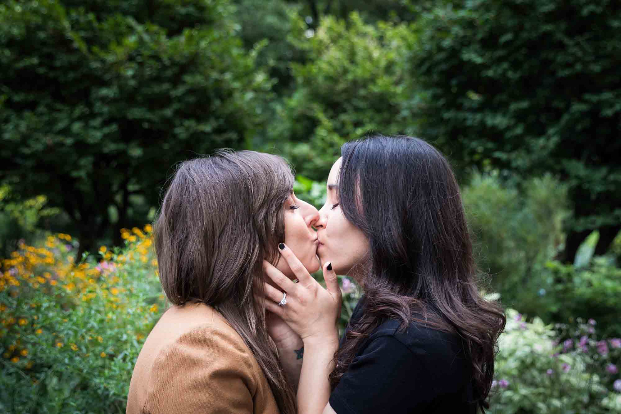 Two women kissing in Riverside Park surrounded by green foliage and flowers for an article on how to produce a movie-themed surprise proposal