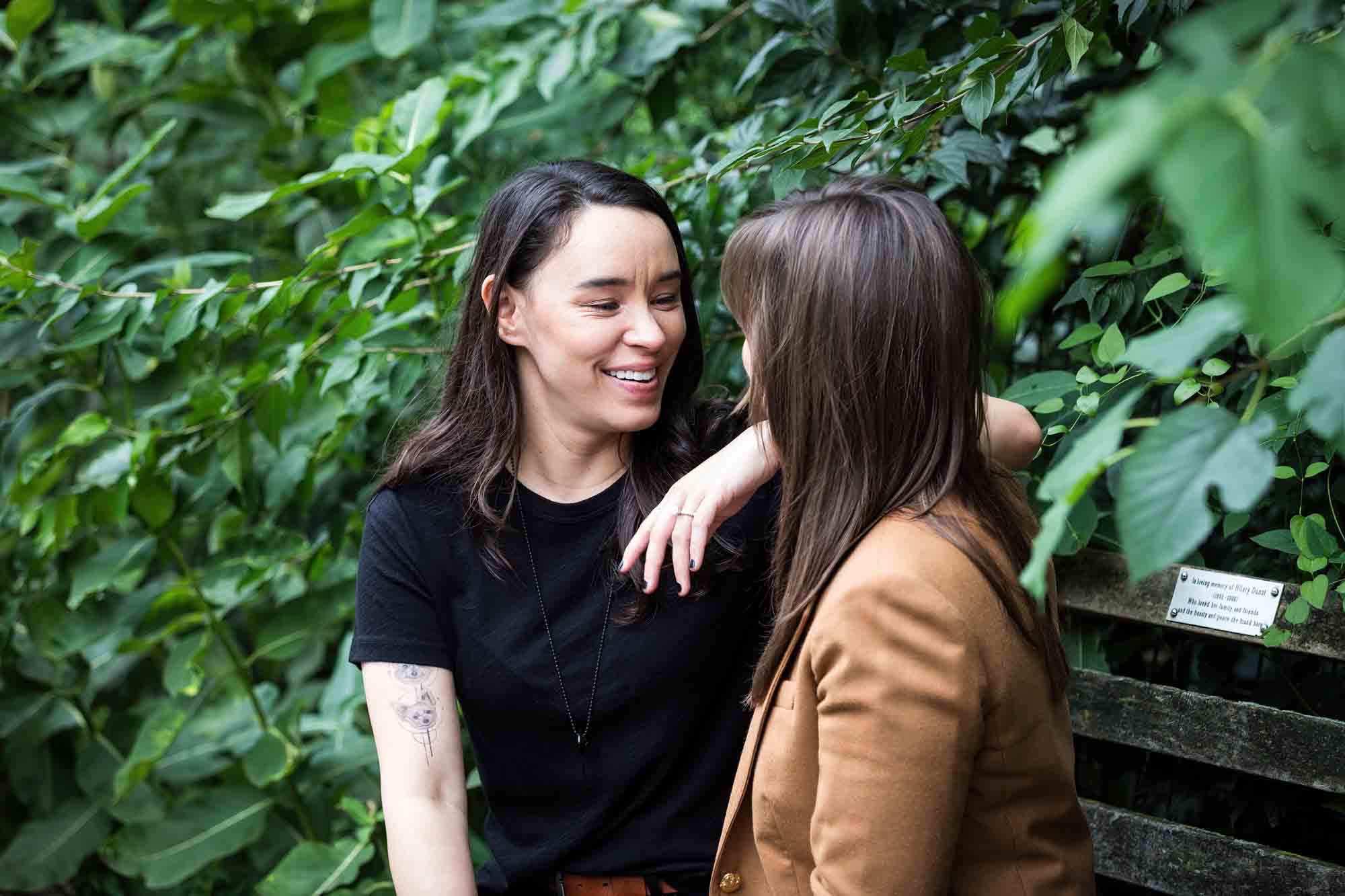 Two women sitting on a bench in front of green foliage in Riverside Park for an article on how to produce a movie-themed surprise proposal