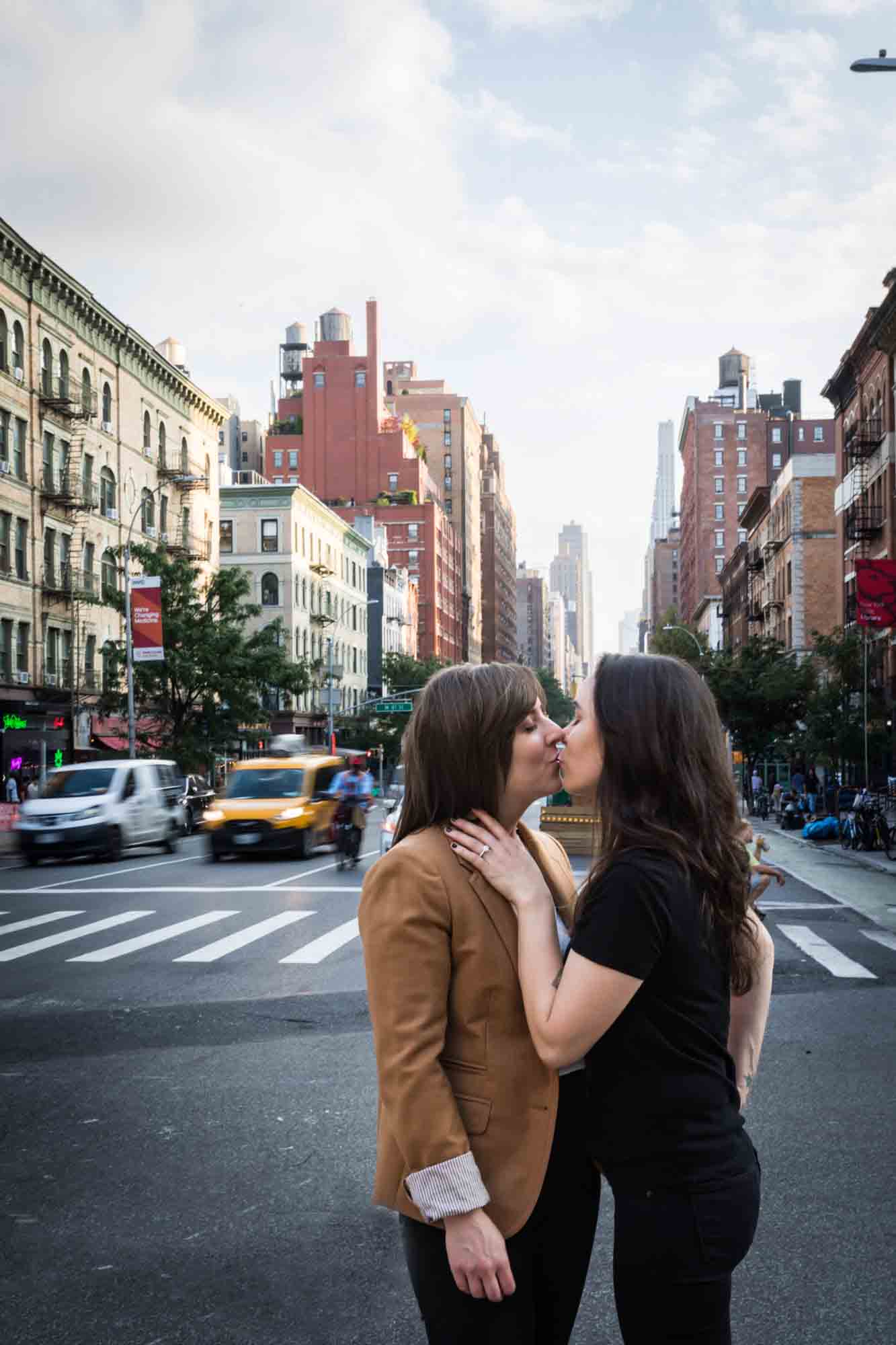 Two women kissing in a NYC roadway with traffic and apartment buildings in the background