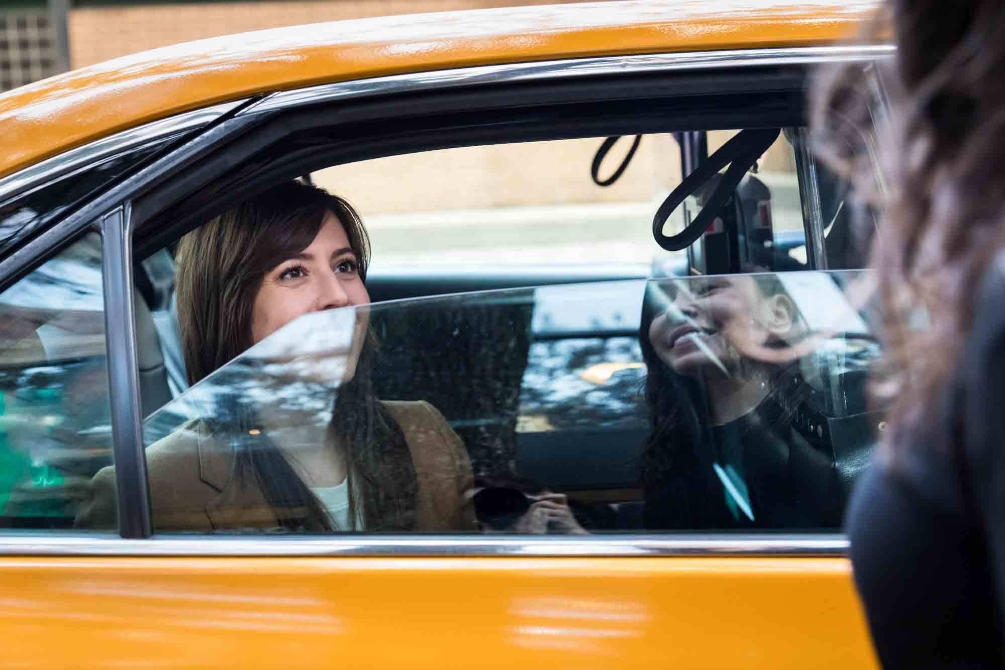 Woman in yellow taxi cab smiling through the window at another woman reflected in the window