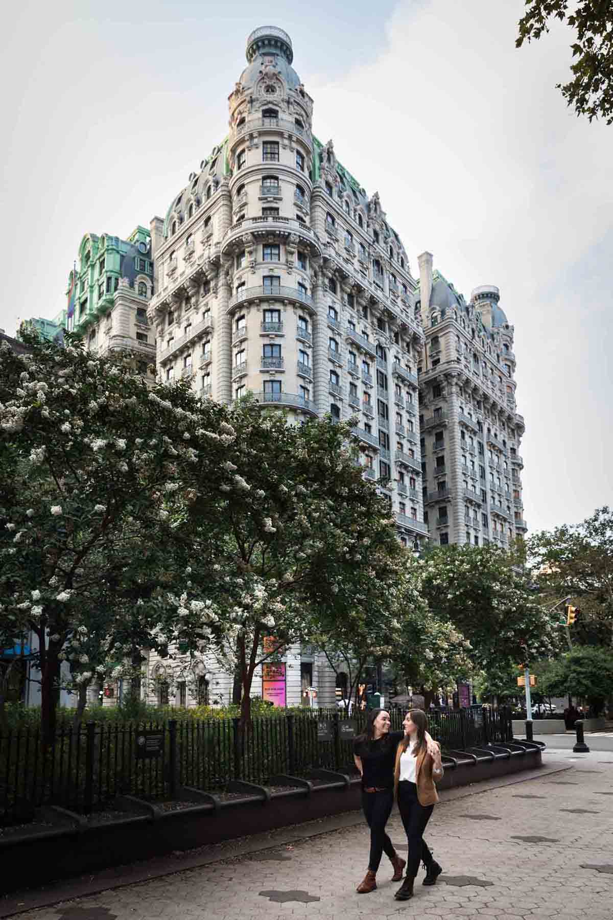 Two women walking through Verdi Square in NYC in front of the Ansonia apartment building