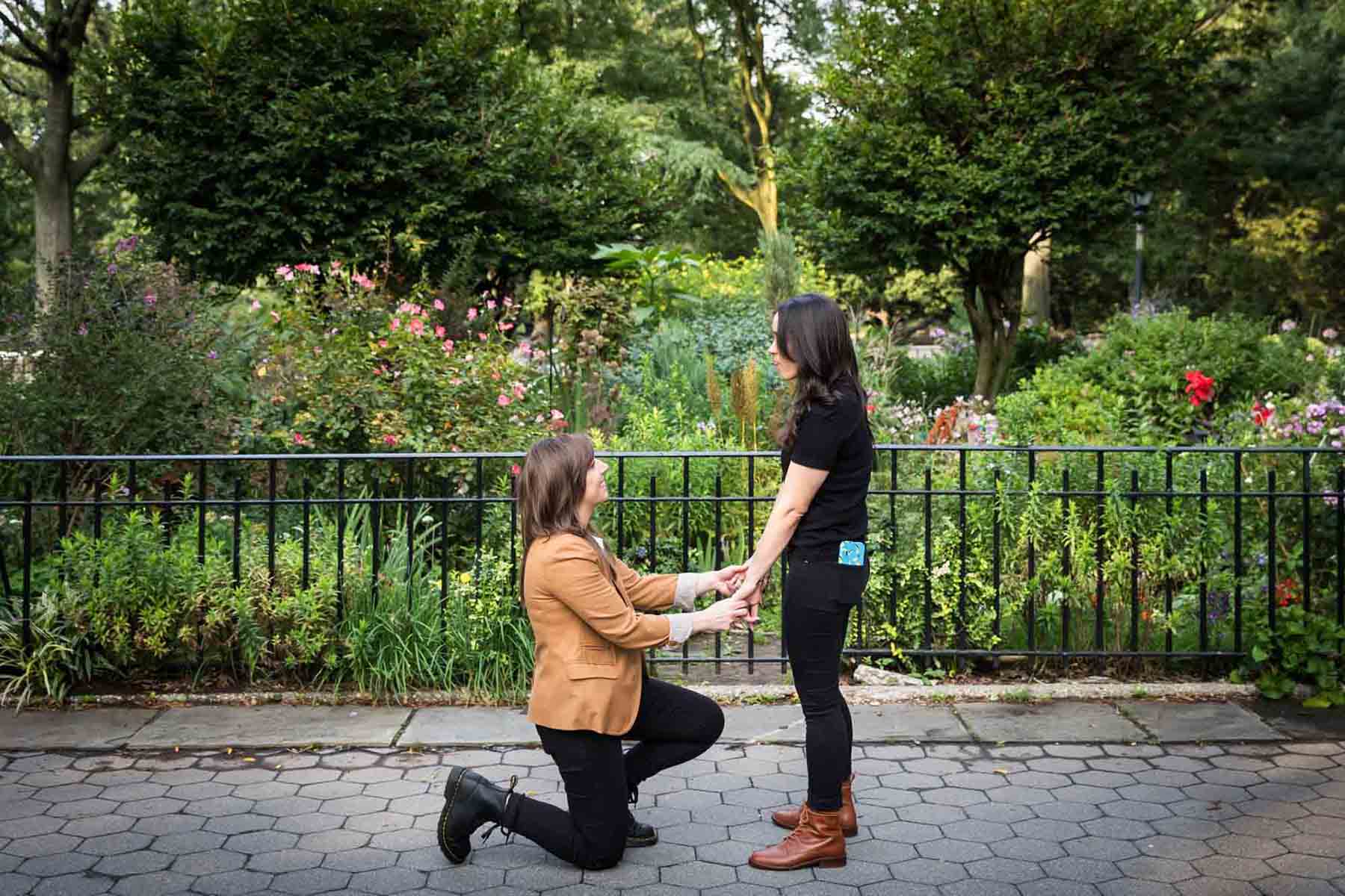 Woman on one knee holding hands with another woman during Riverside Park surprise proposal
