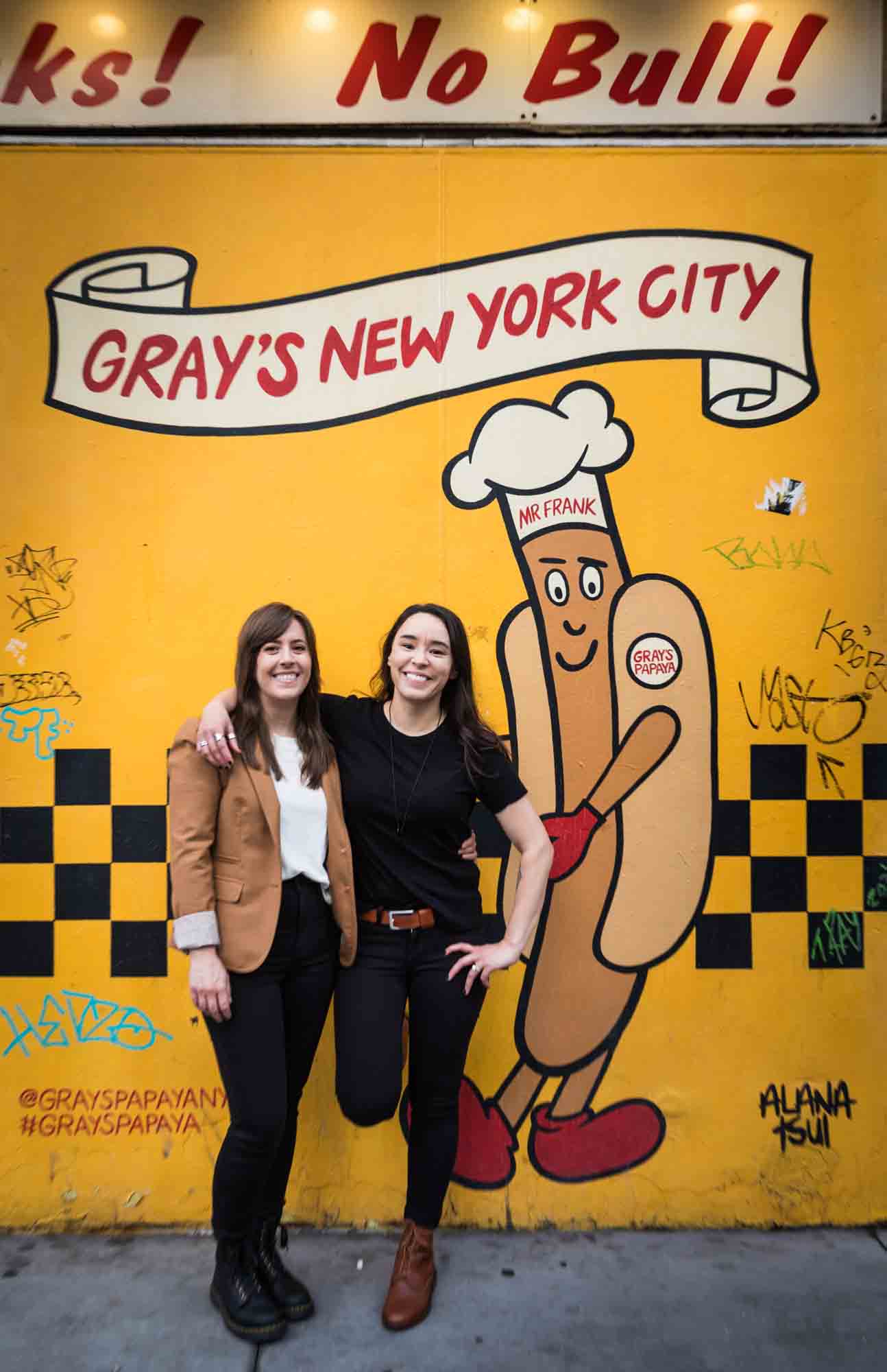 Two women hugging in front of the image of a cartoon hot dog character