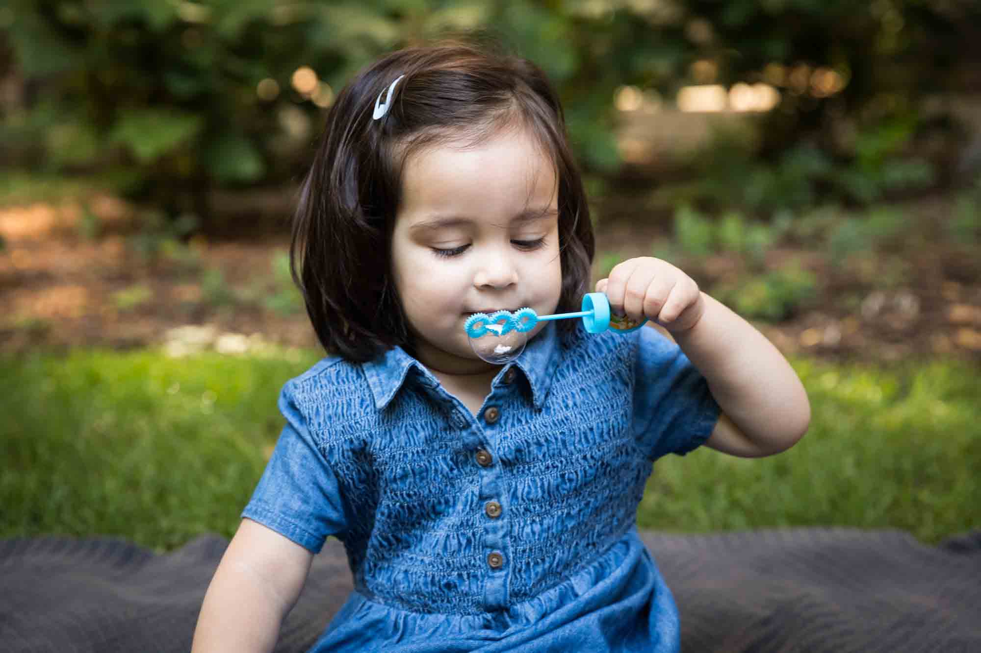 Little girl in blue dress blowing bubbles in grass during a Brooklyn Commons family portrait session
