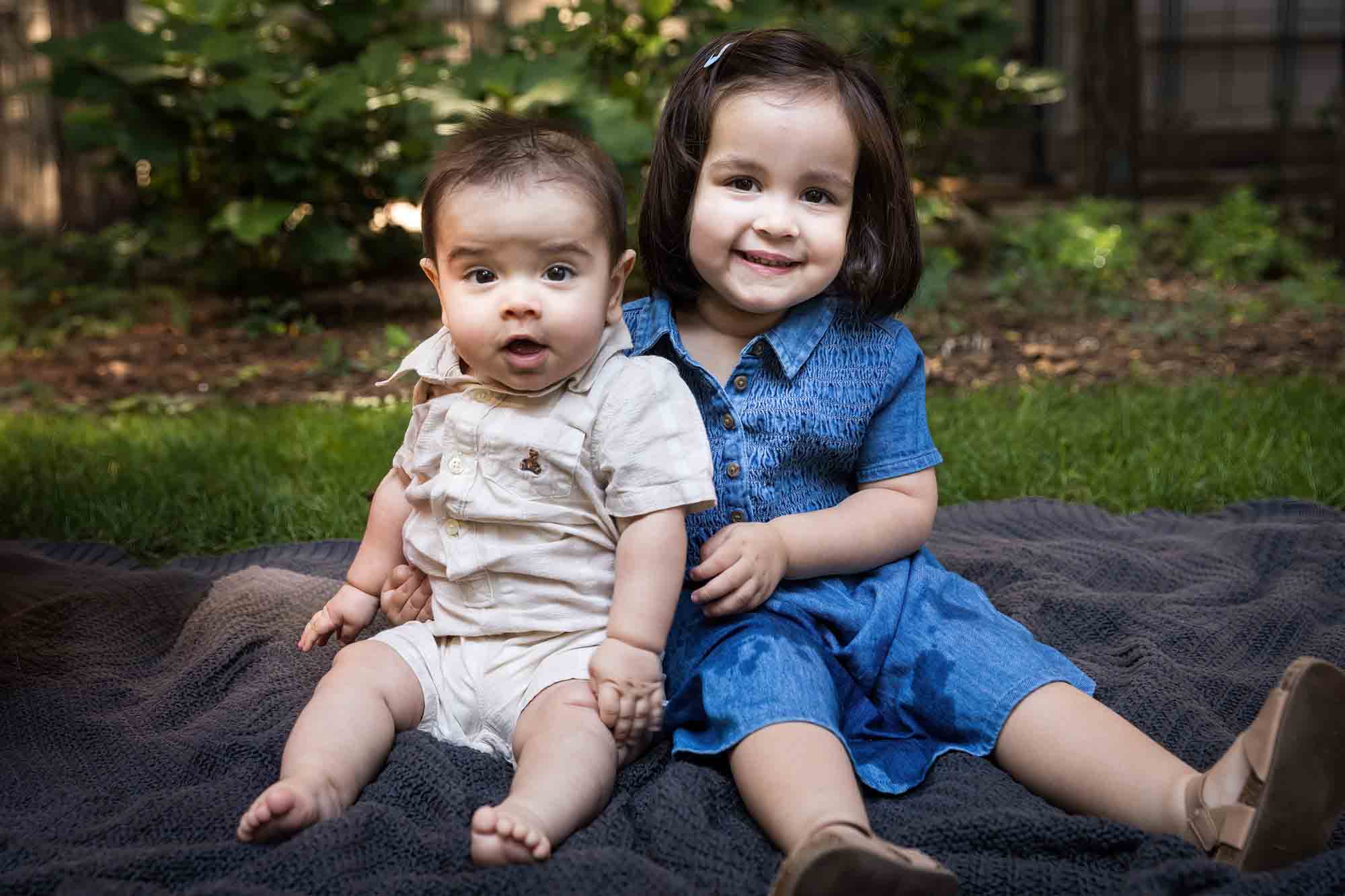 Little girl in blue dress holding baby brother on gray blanket during a Brooklyn Commons family portrait session