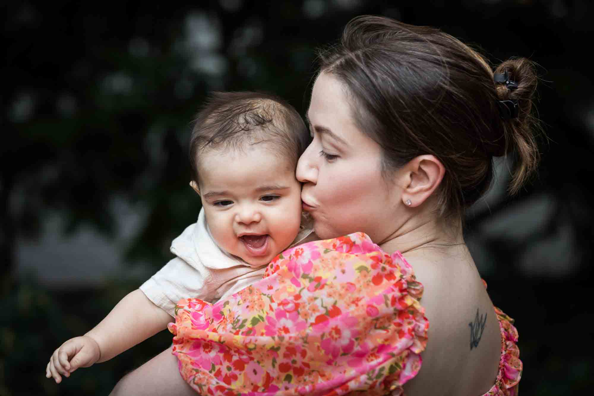 Mother in red floral dress kissing baby boy during a Brooklyn Commons family portrait session