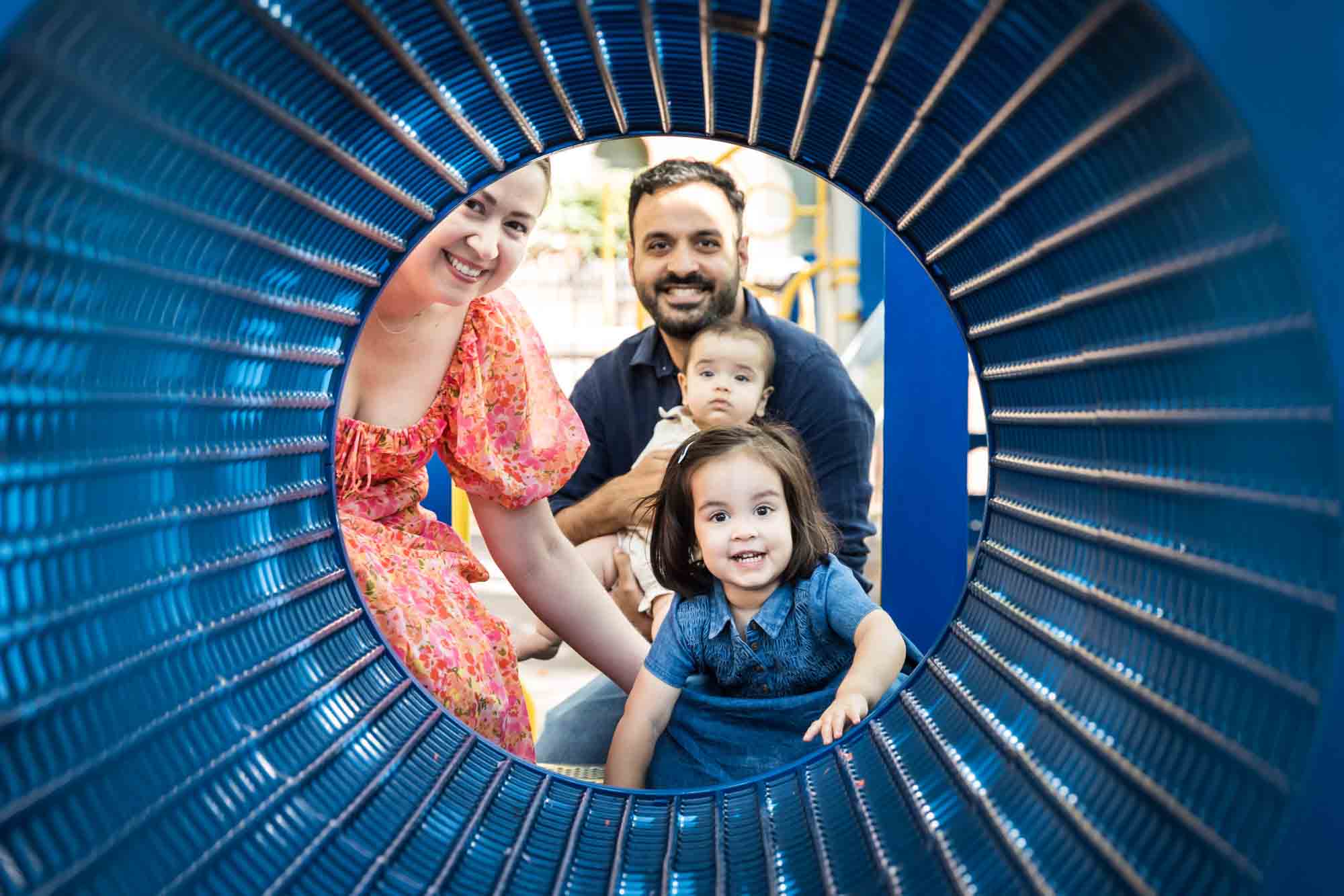 Parents with little girl and baby boy seen through blue playground tunnel