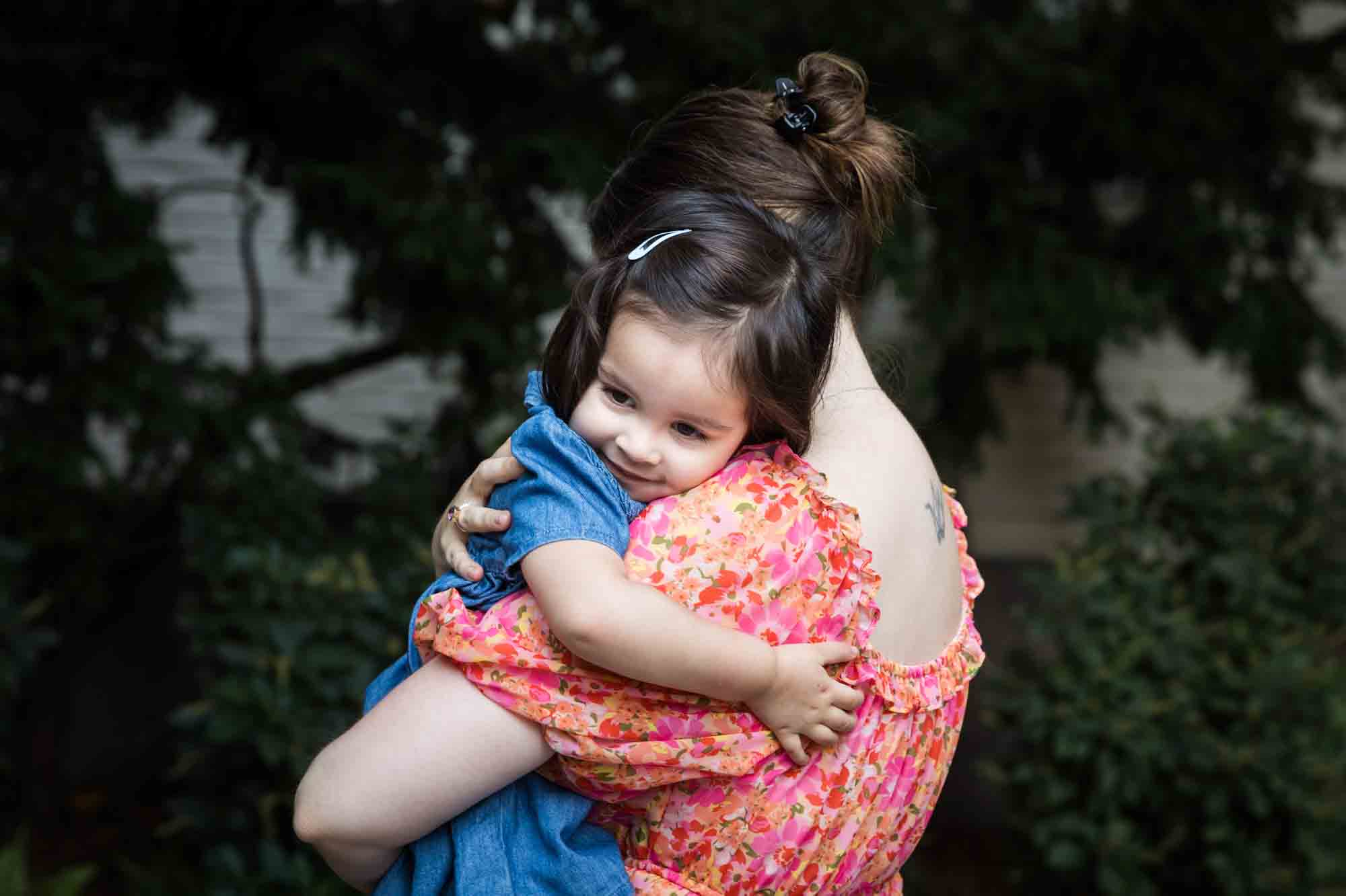 Mother in floral dress holding little girl in blue dress in front of vine-covered wall during a Brooklyn Commons family portrait session