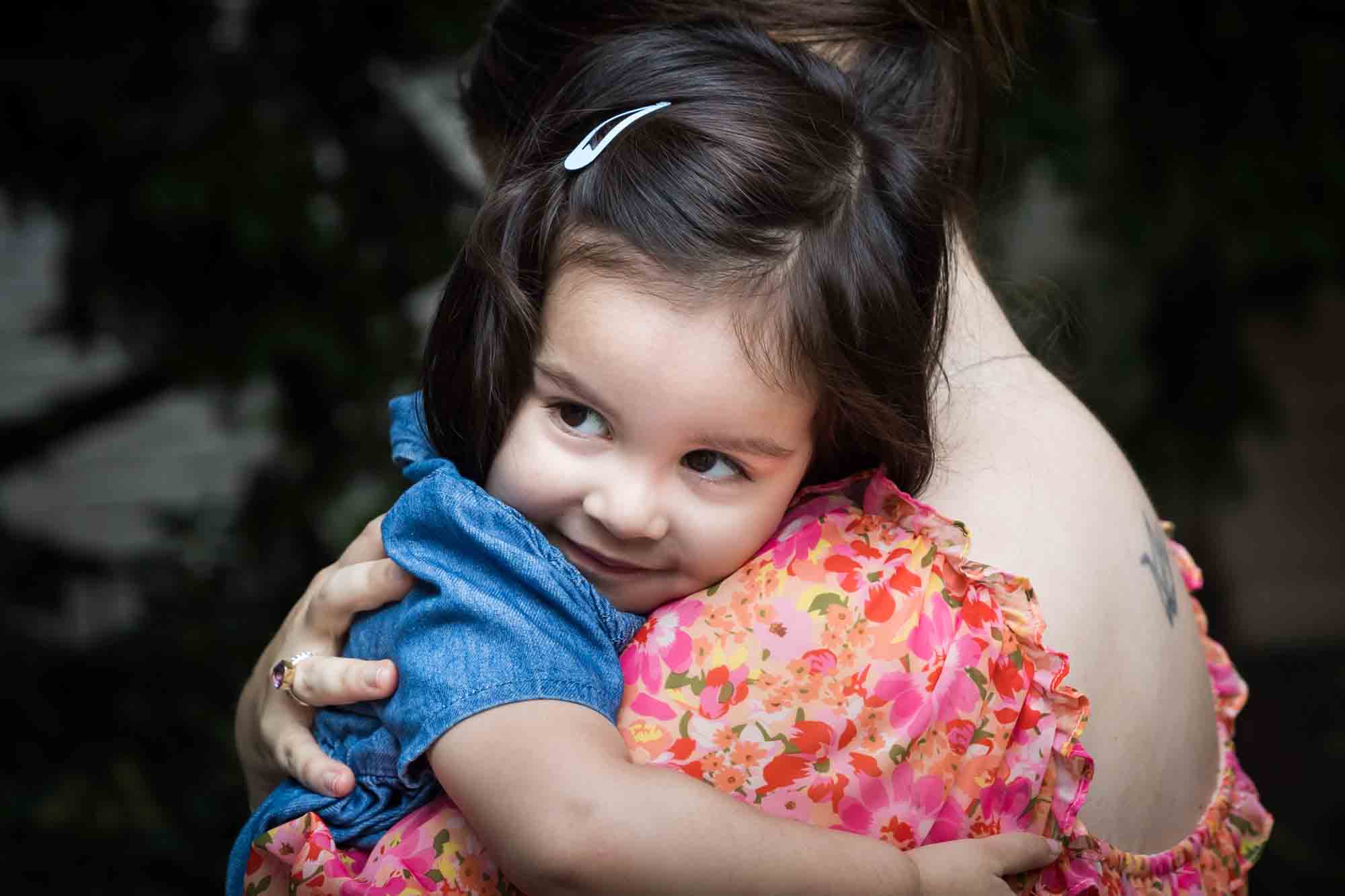 Mother in floral dress holding little girl in blue dress in front of vine-covered wall during a Brooklyn Commons family portrait session