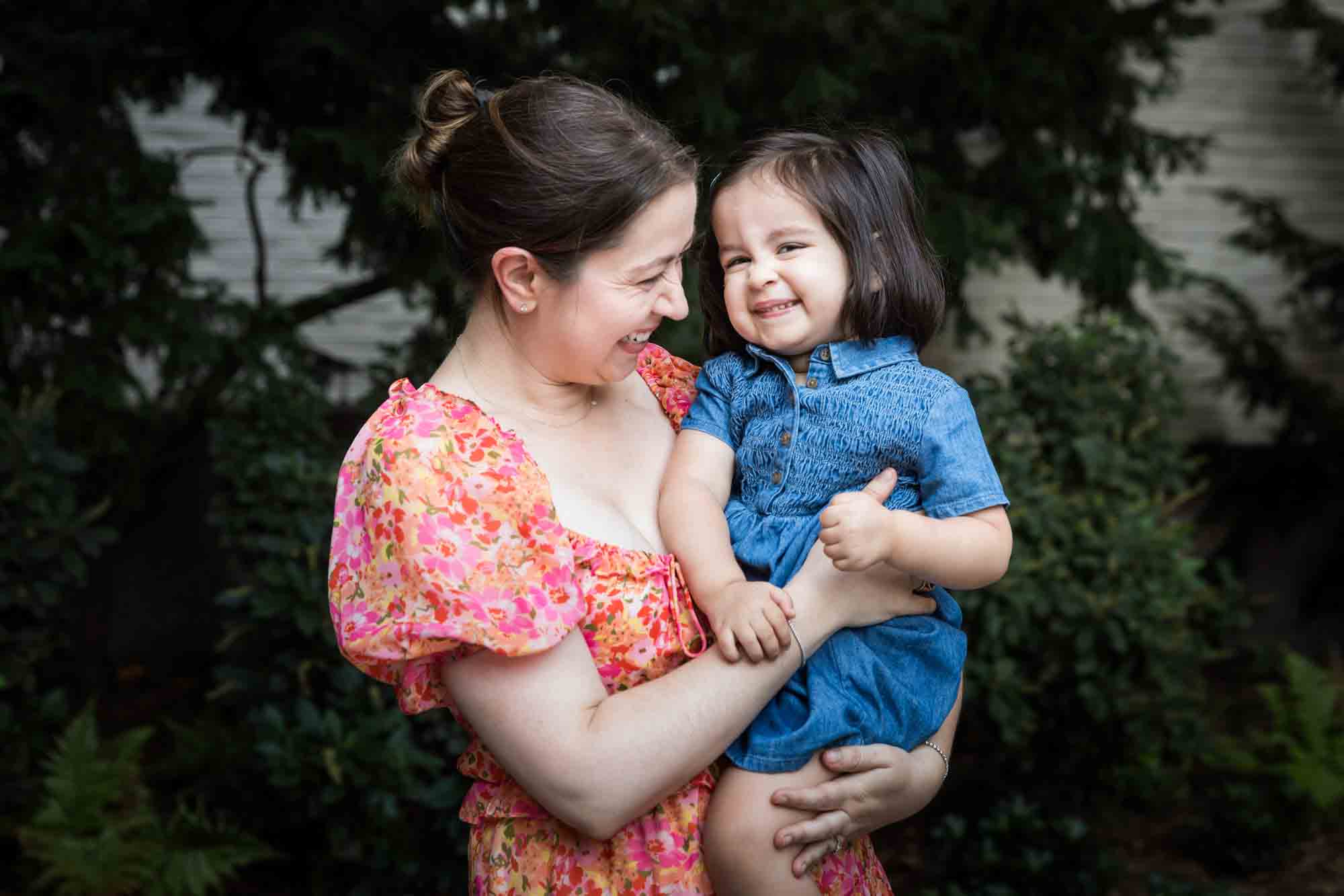 Mother in floral dress holding little girl in blue dress in front of vine-covered wall during a Brooklyn Commons family portrait session