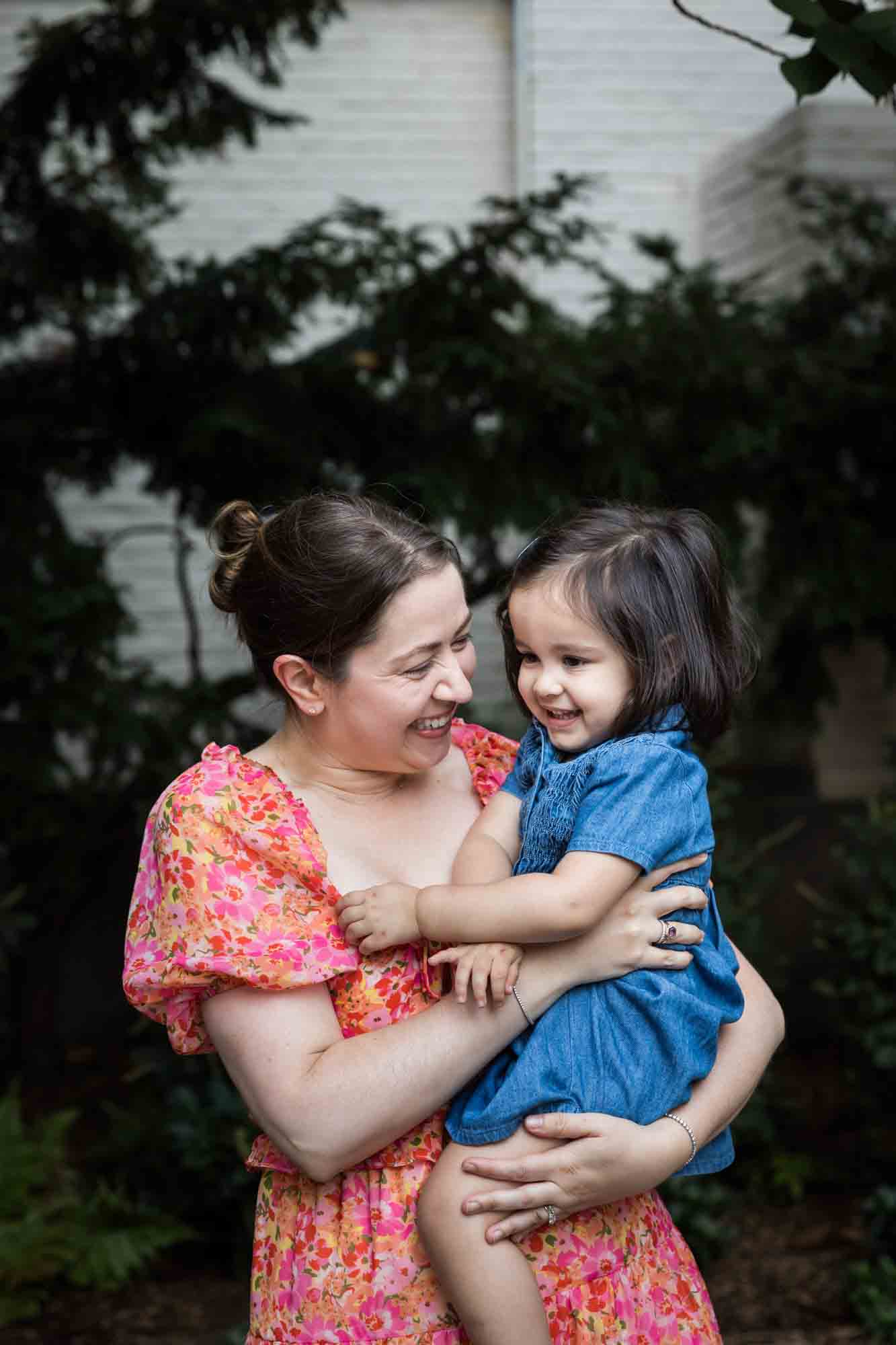 Mother in floral dress holding little girl in blue dress in front of vine-covered wall during a Brooklyn Commons family portrait session