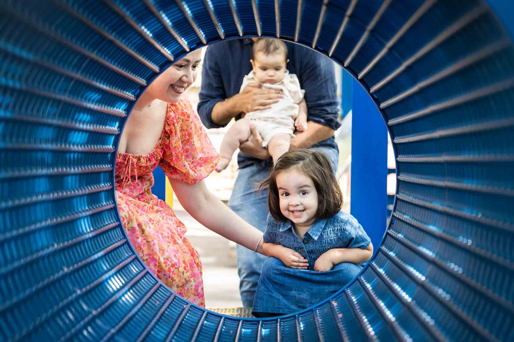 Parents with little girl and baby boy seen through blue playground tunnel