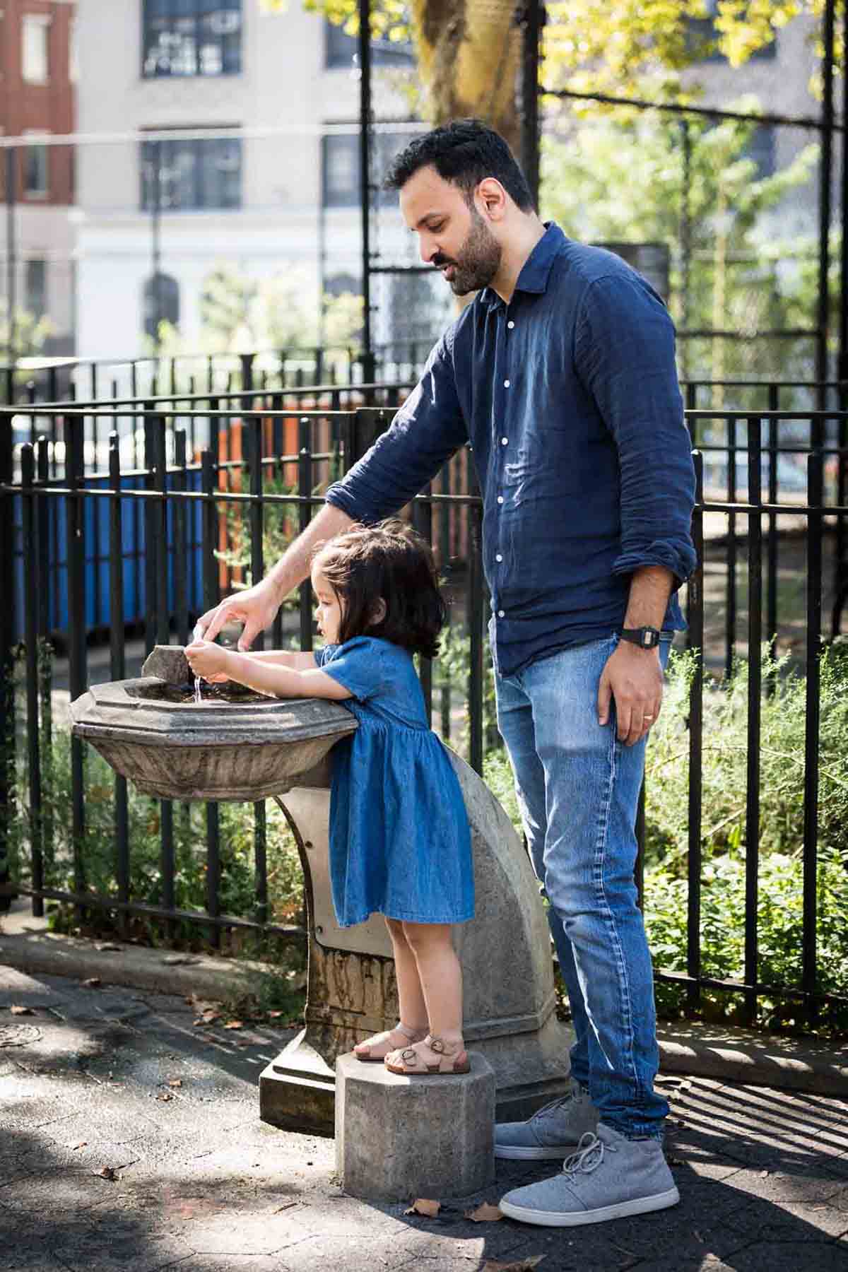 Father helping little girl in blue dress wash hands in water fountain in McLaughlin Park