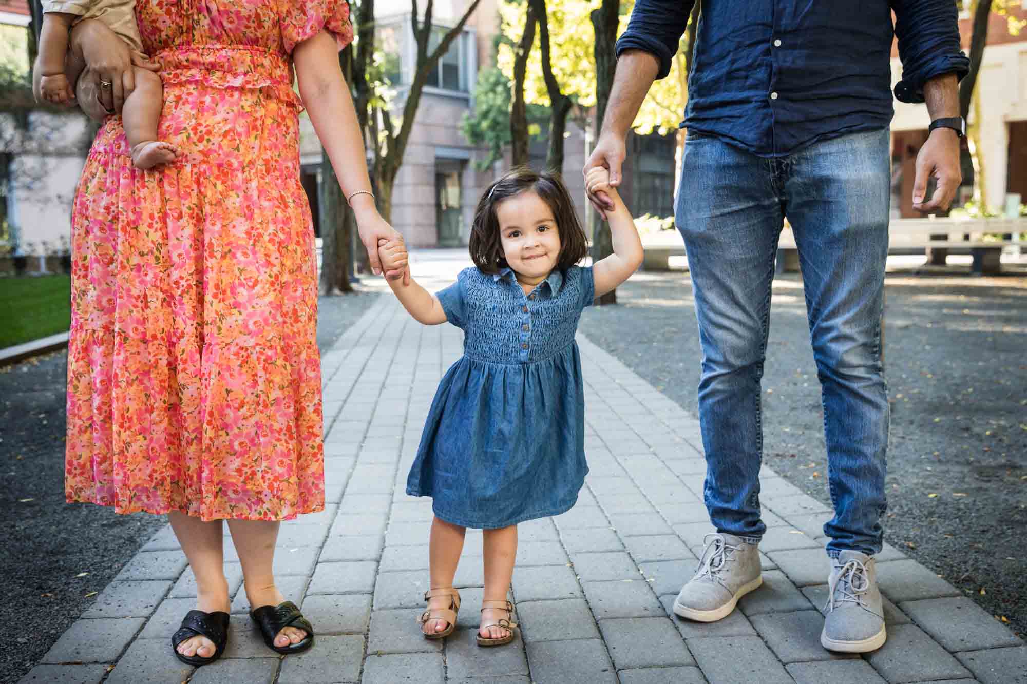 Little girl in blue dress holding parents hands on stone pathway during a Brooklyn Commons family portrait session