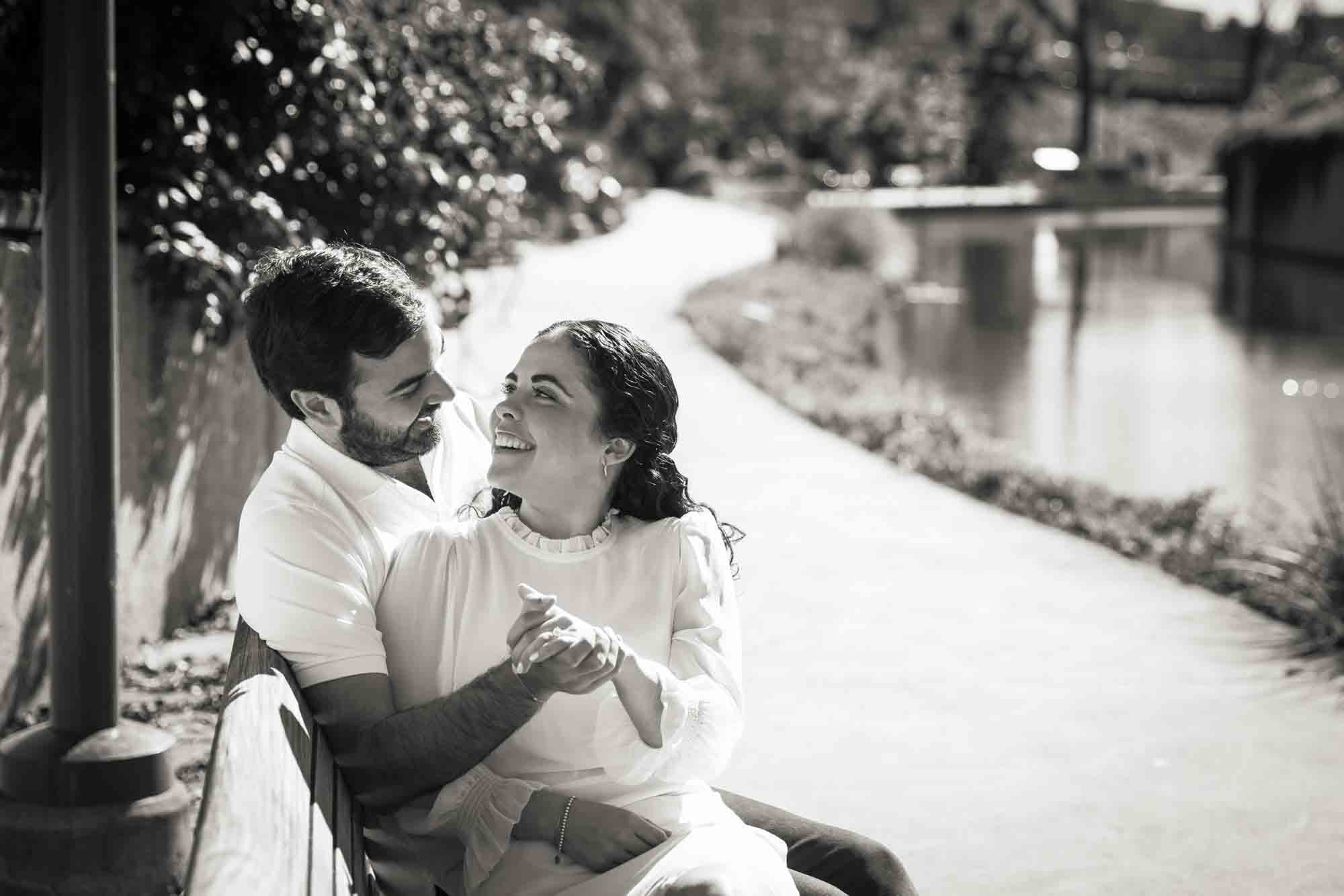 Black and white photo of couple sitting on bench beside the Riverwalk during a Pearl engagement portrait session