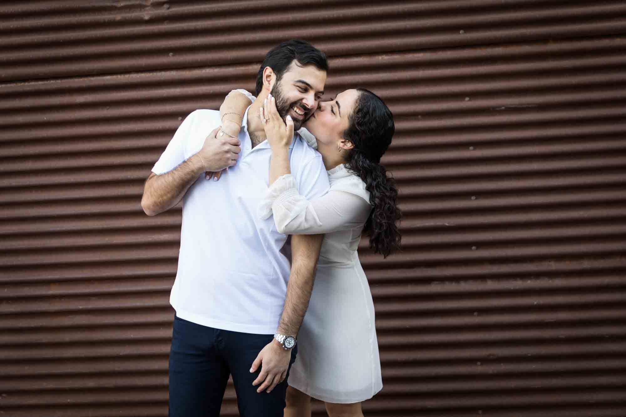 Woman kissing man on the cheek in front of rusty metal door during a Pearl engagement portrait session