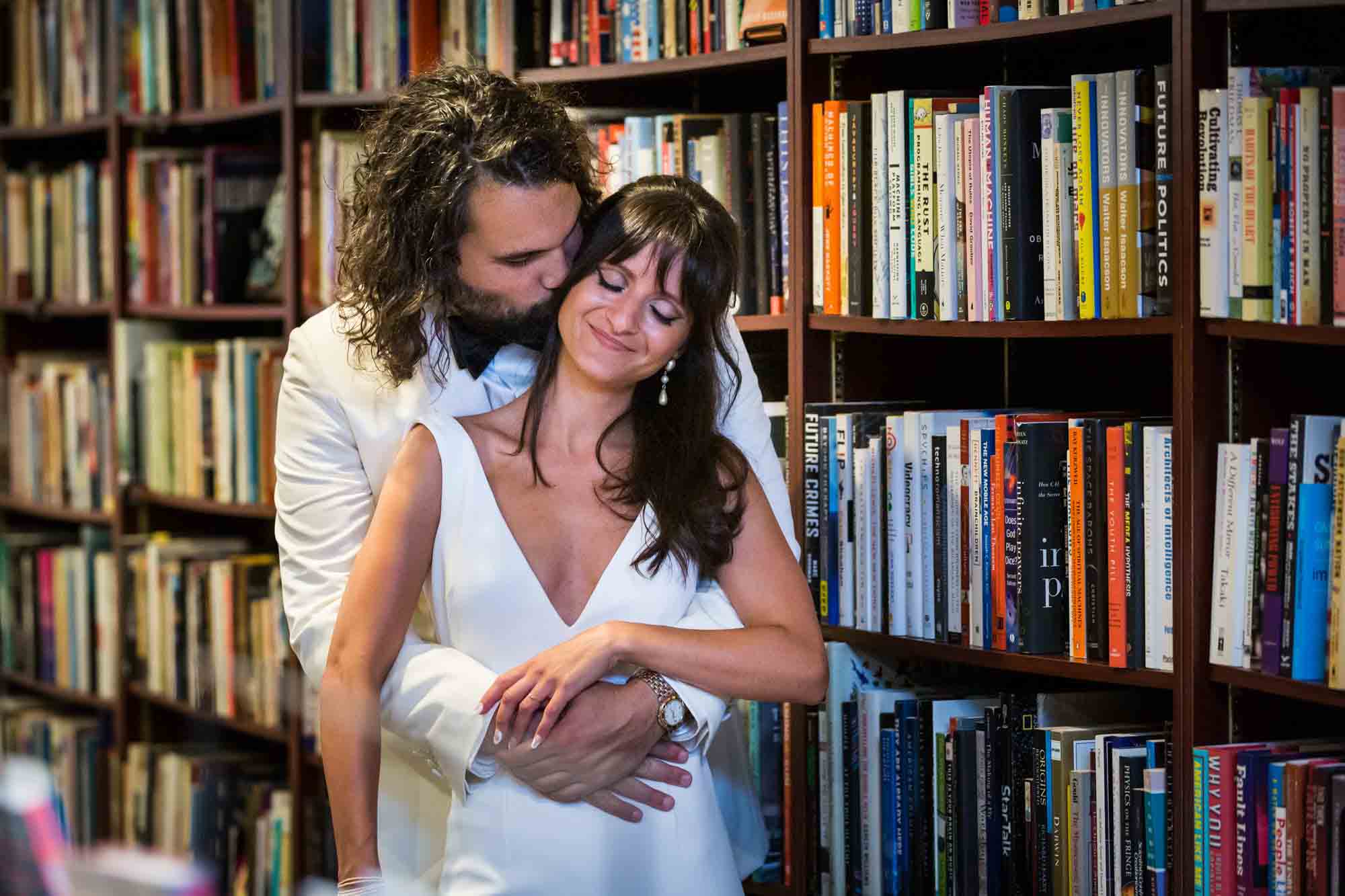 Groom hugging bride from behind in front of bookshelf