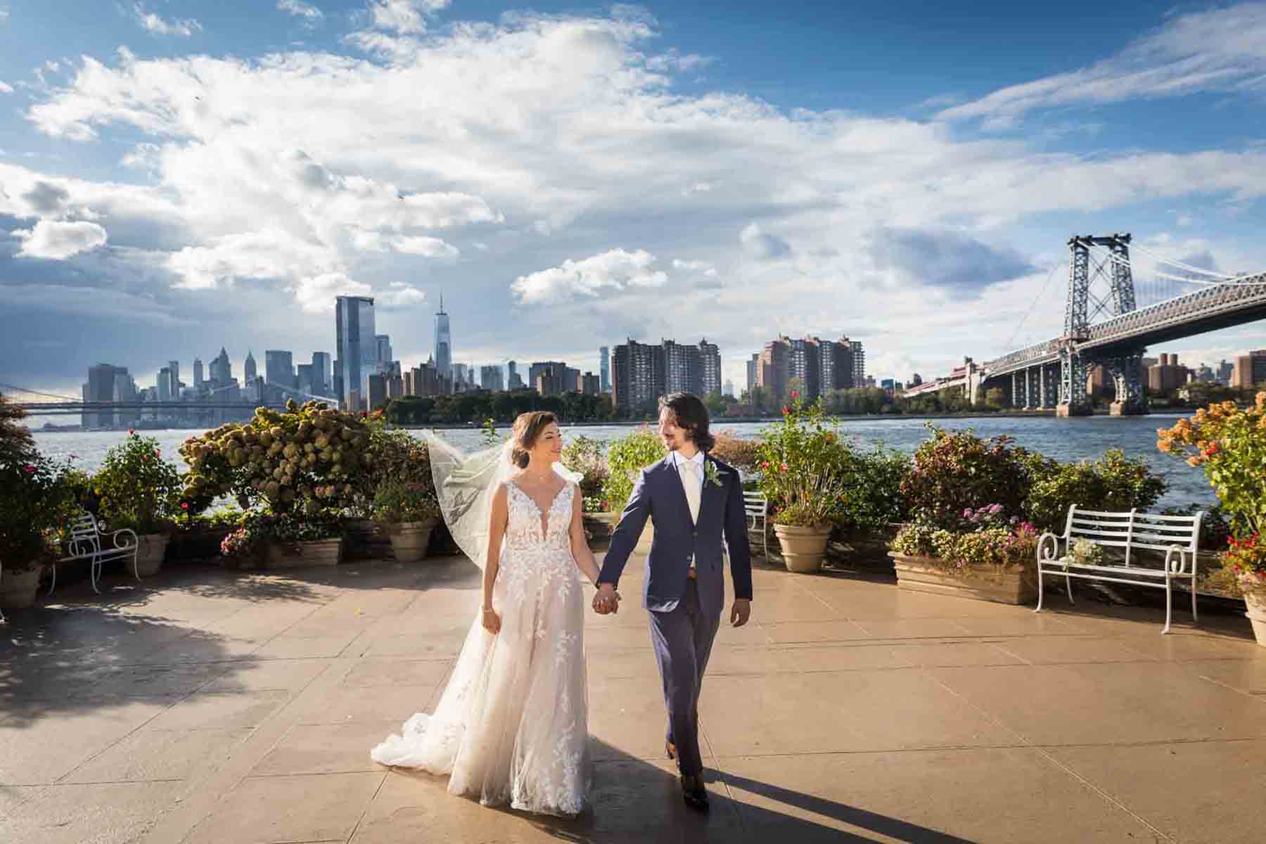 Bride and groom holding hands and walking on patio with NYC skyline in background