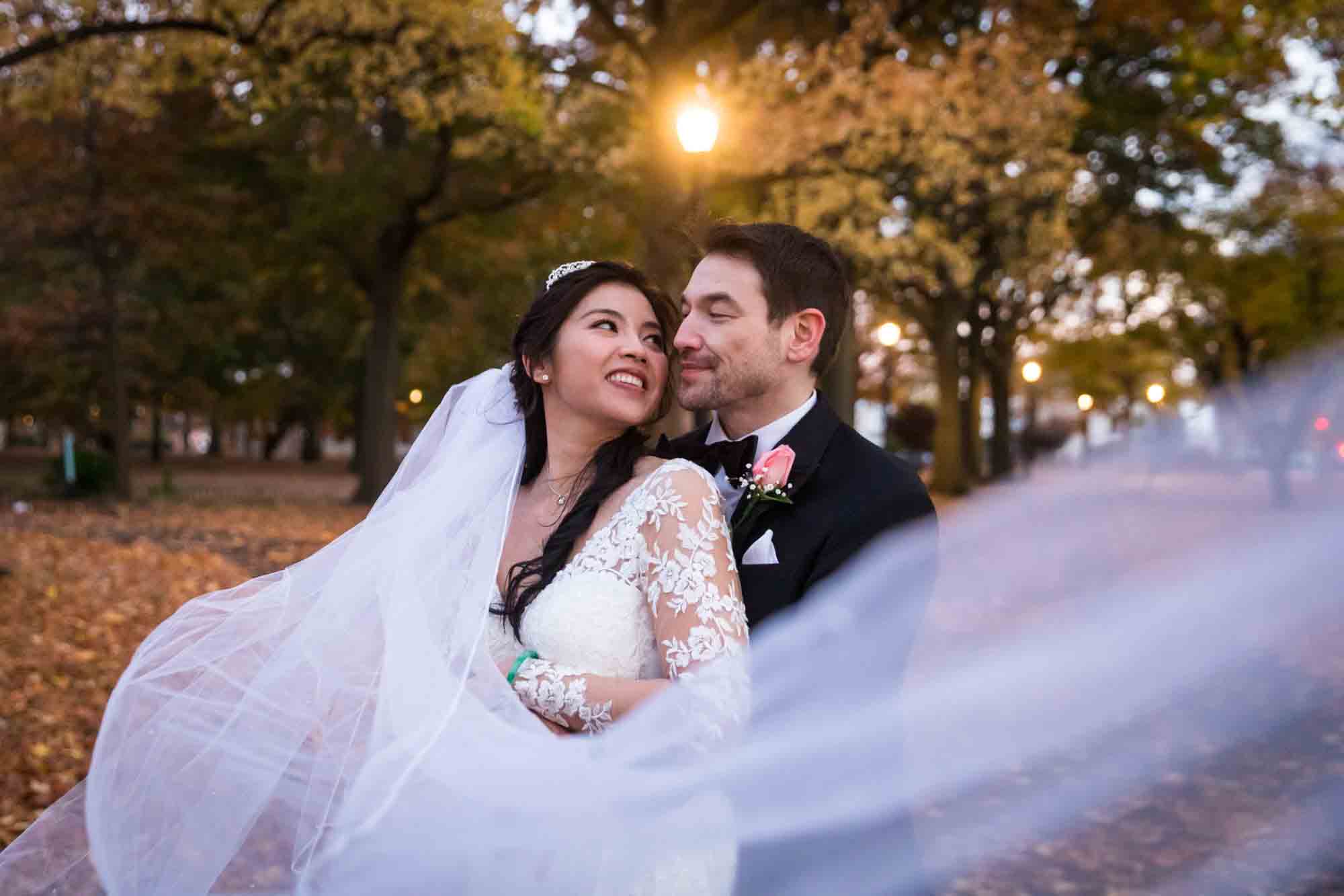 Bride and groom hugging behind veil at dusk with fall trees behind