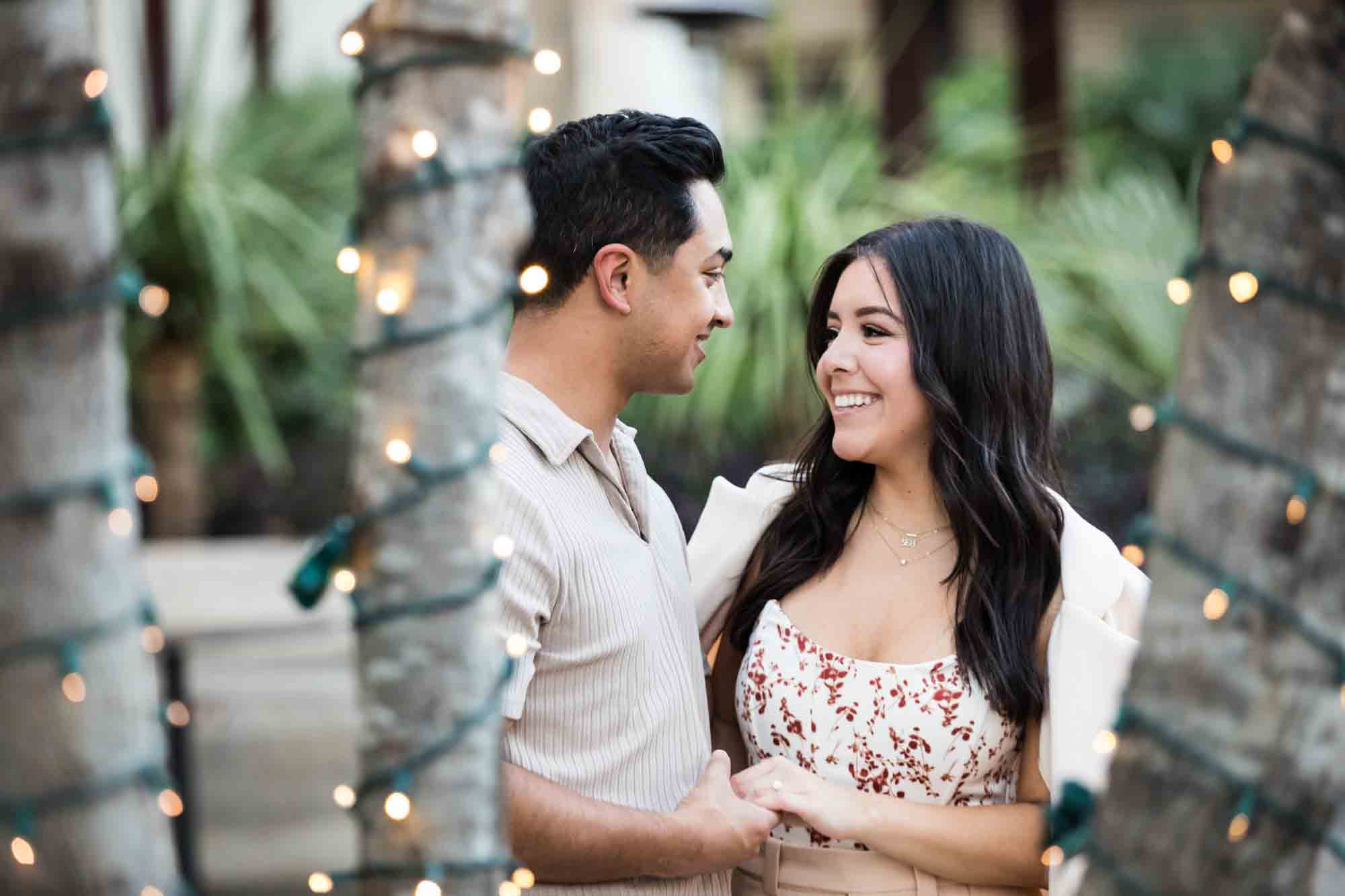 Couple holding hands and smiling at each other in between trees covered in fairy lights for an article on how to propose at the Pearl