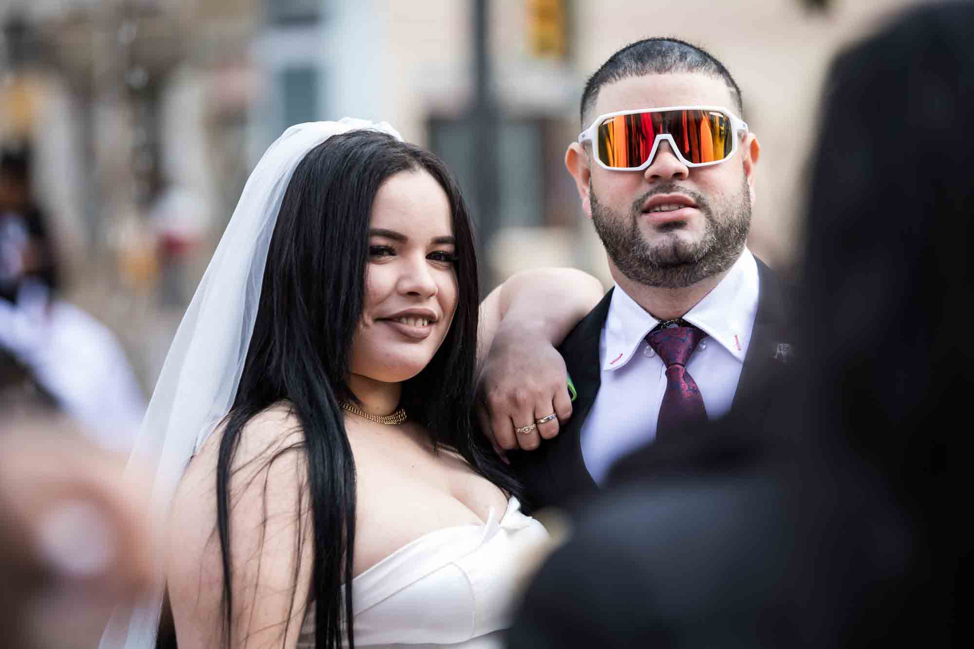Bride hanging on shoulder of groom wearing large reflective sunglasses for an article on the Bexar county mass wedding event