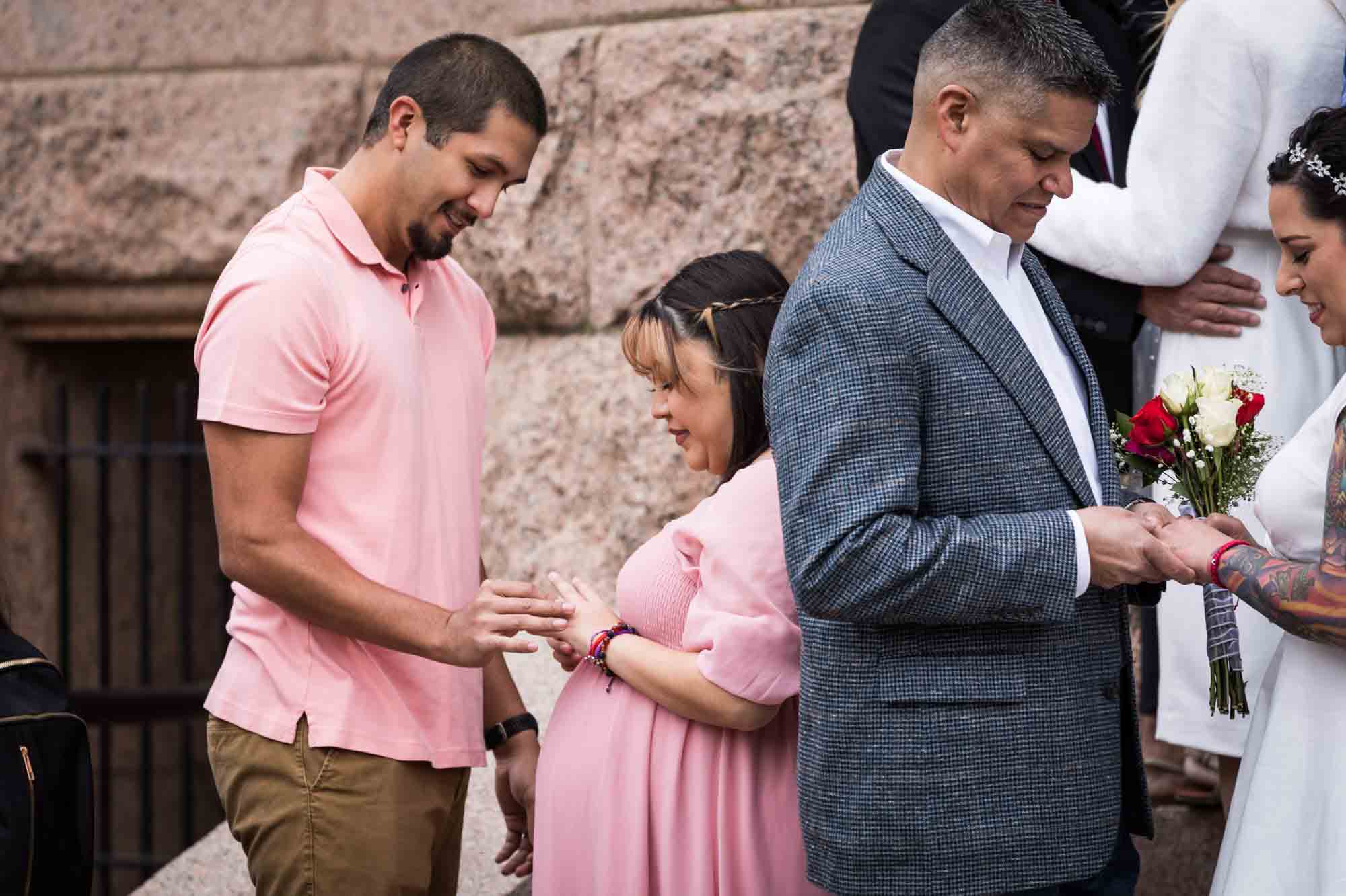 Bride in pink dress putting ring on hand of groom wearing pink shirt 