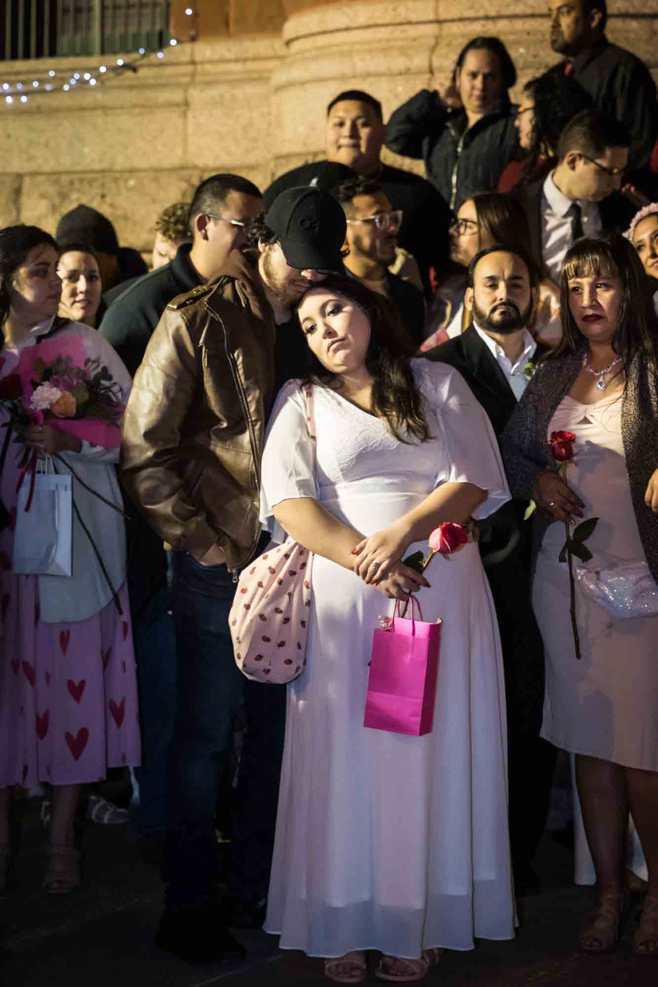 Bride in white dress leaning on man wearing black cap in front of crowd for an article on the Bexar county mass wedding event