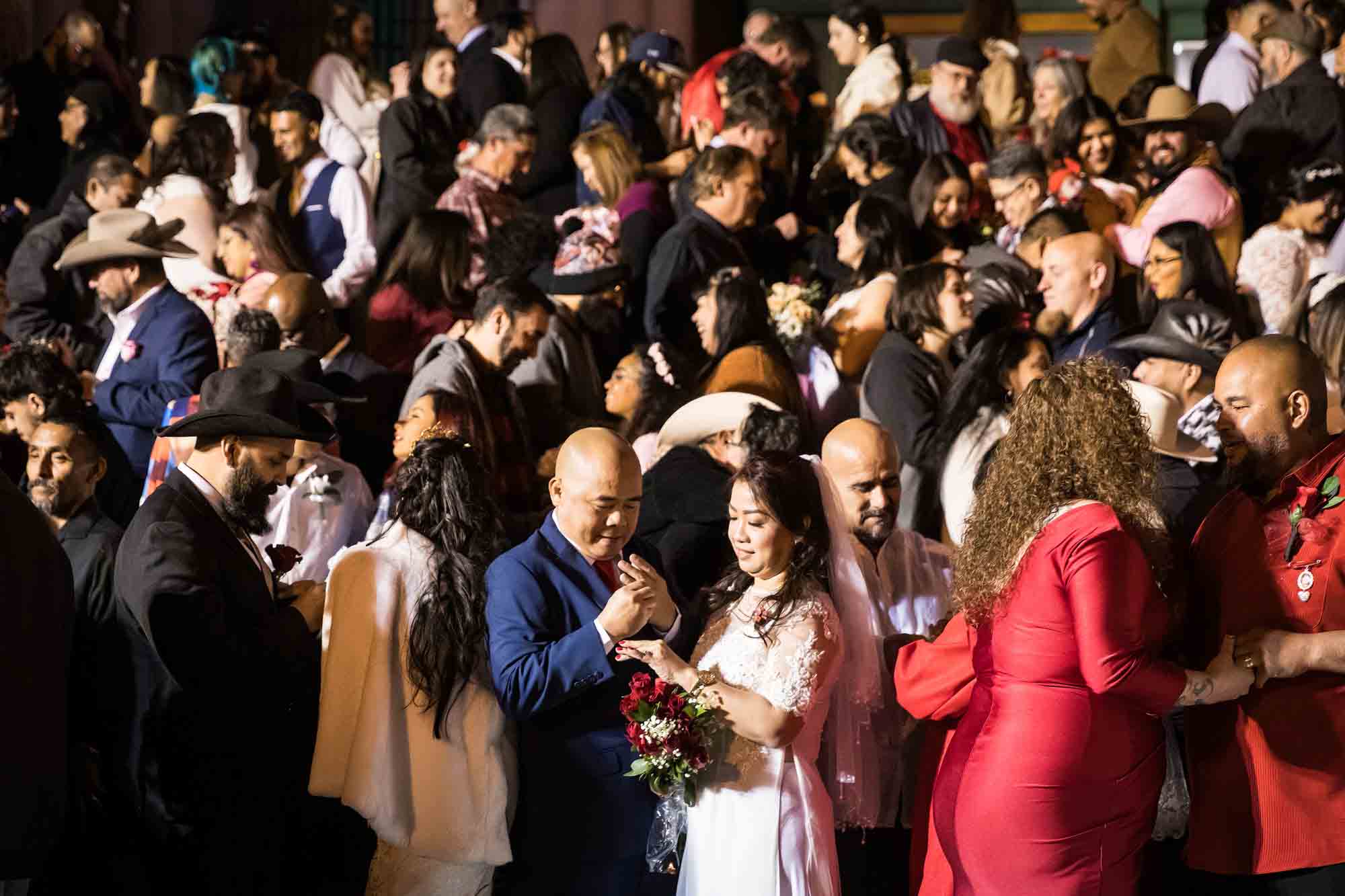 Crowd of bride and grooms putting on rings in front of courthouse steps at night for an article on the Bexar county mass wedding event