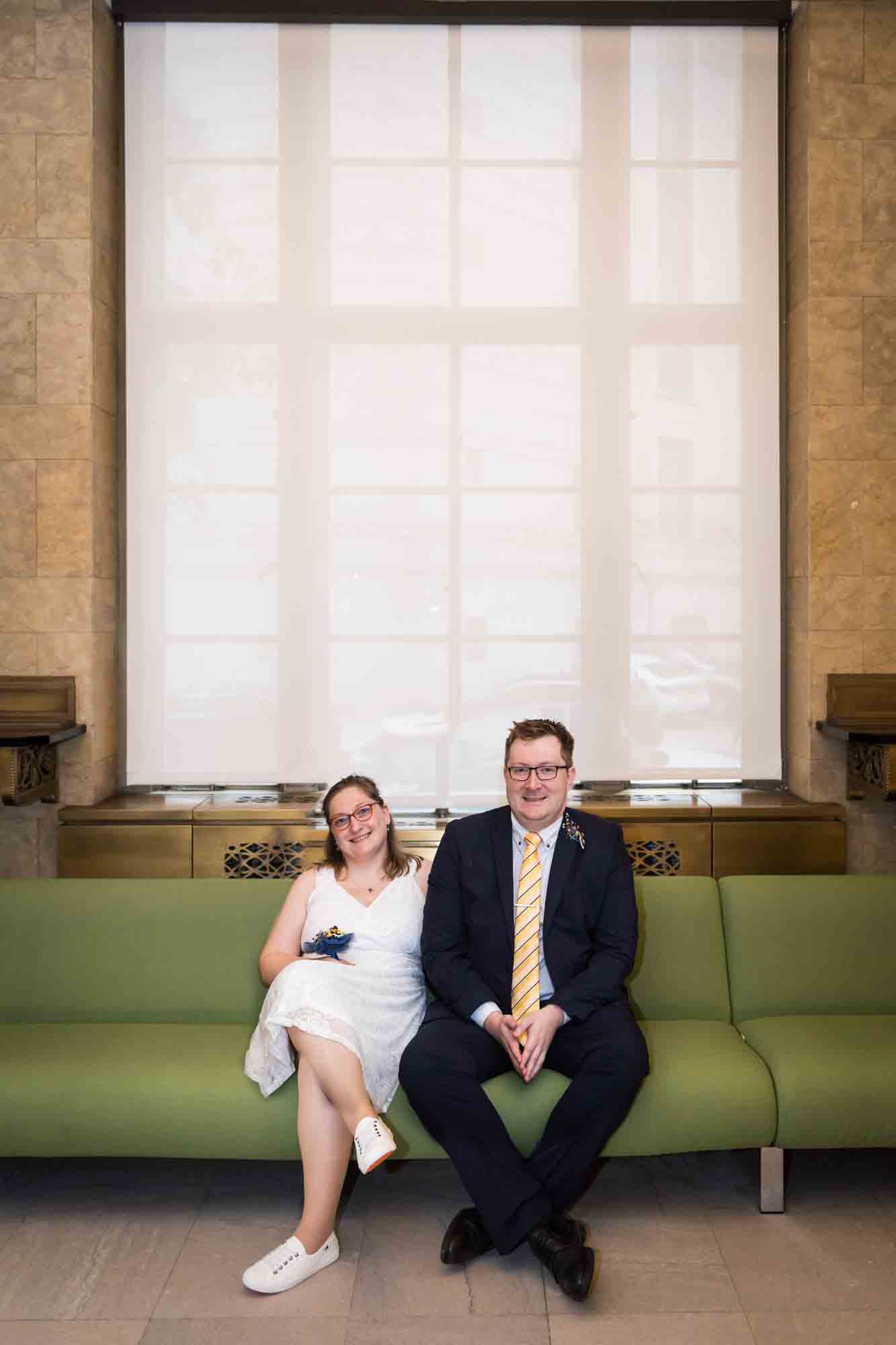 Bride and groom sitting on green couch in front of large window with white shade for an article on how to get a marriage license in San Antonio