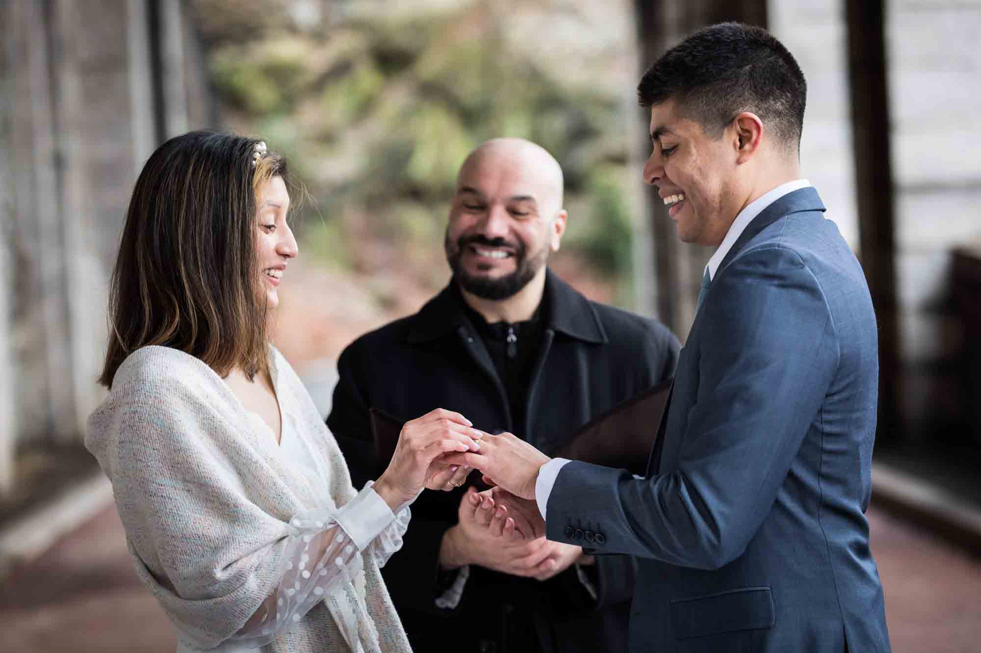 Bride putting wedding ring on groom's finger in front of officiant