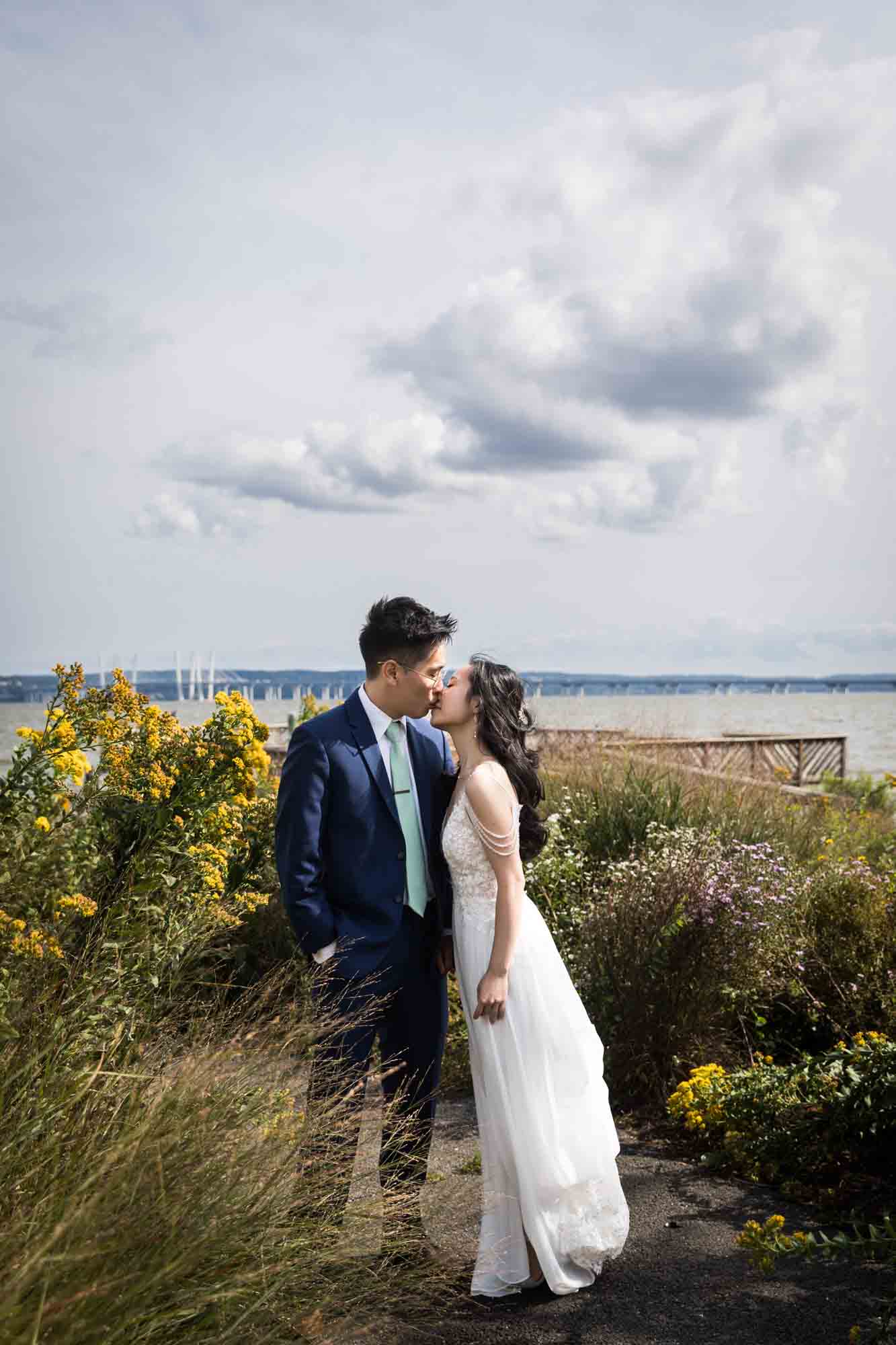 Bride and groom kissing in front of bushes with sky and clouds in background