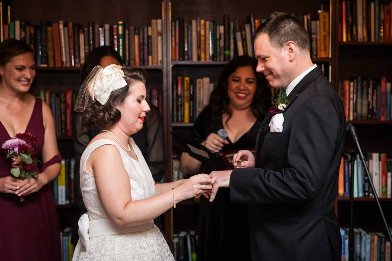 Bride putting ring on groom's finger in front of bookcase for an article on how to become a wedding officiant in San Antonio
