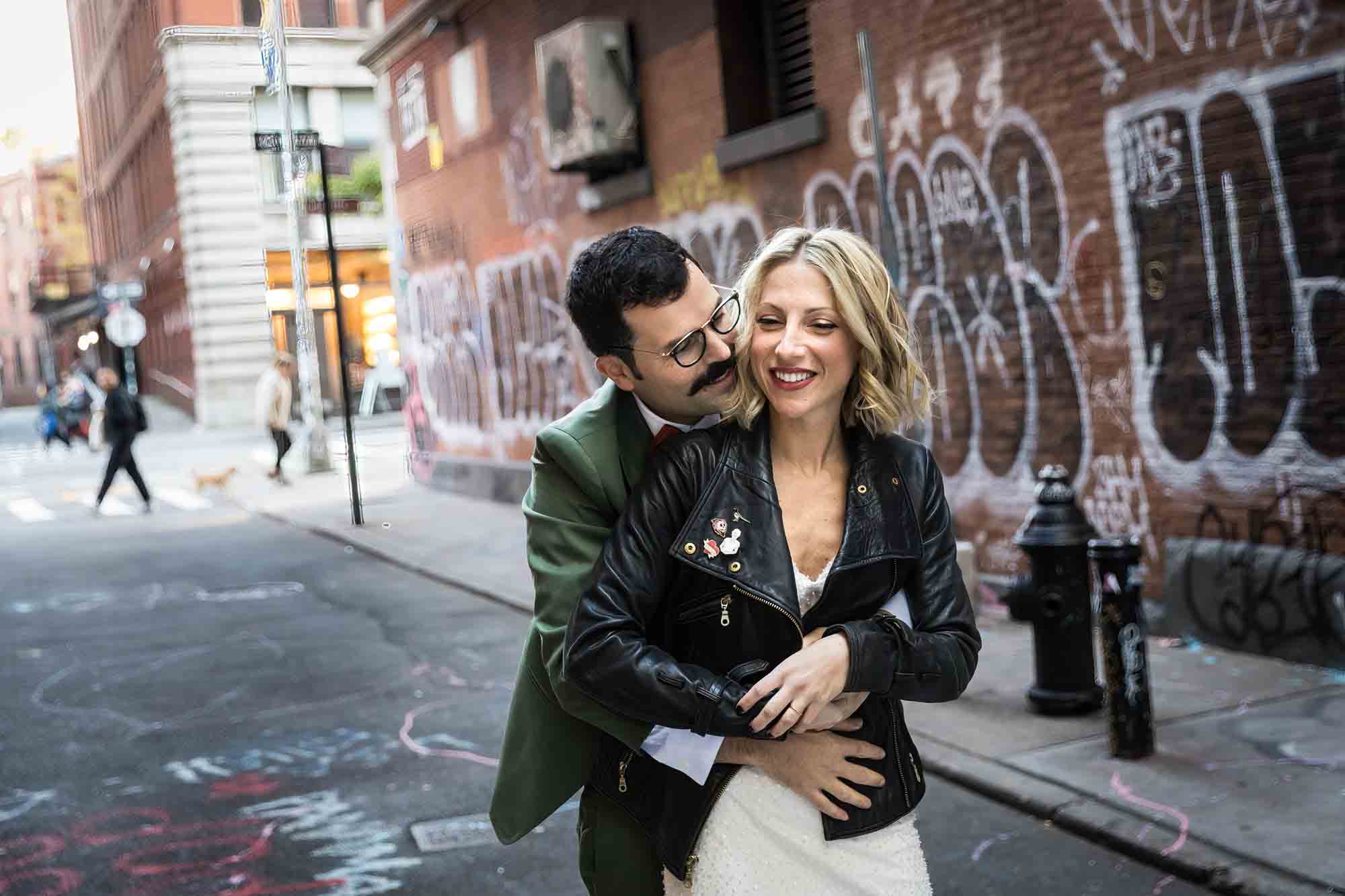 Groom hugging bride from behind in graffiti-filled alleyway for an article on should you give your wedding photographer a shot list