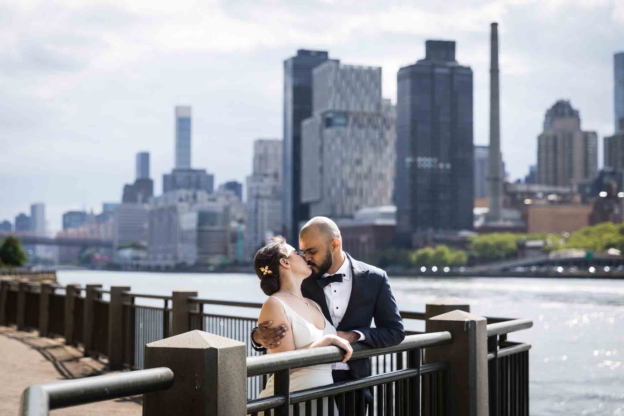 Bride and groom kissing along railing with river and city skyline in background