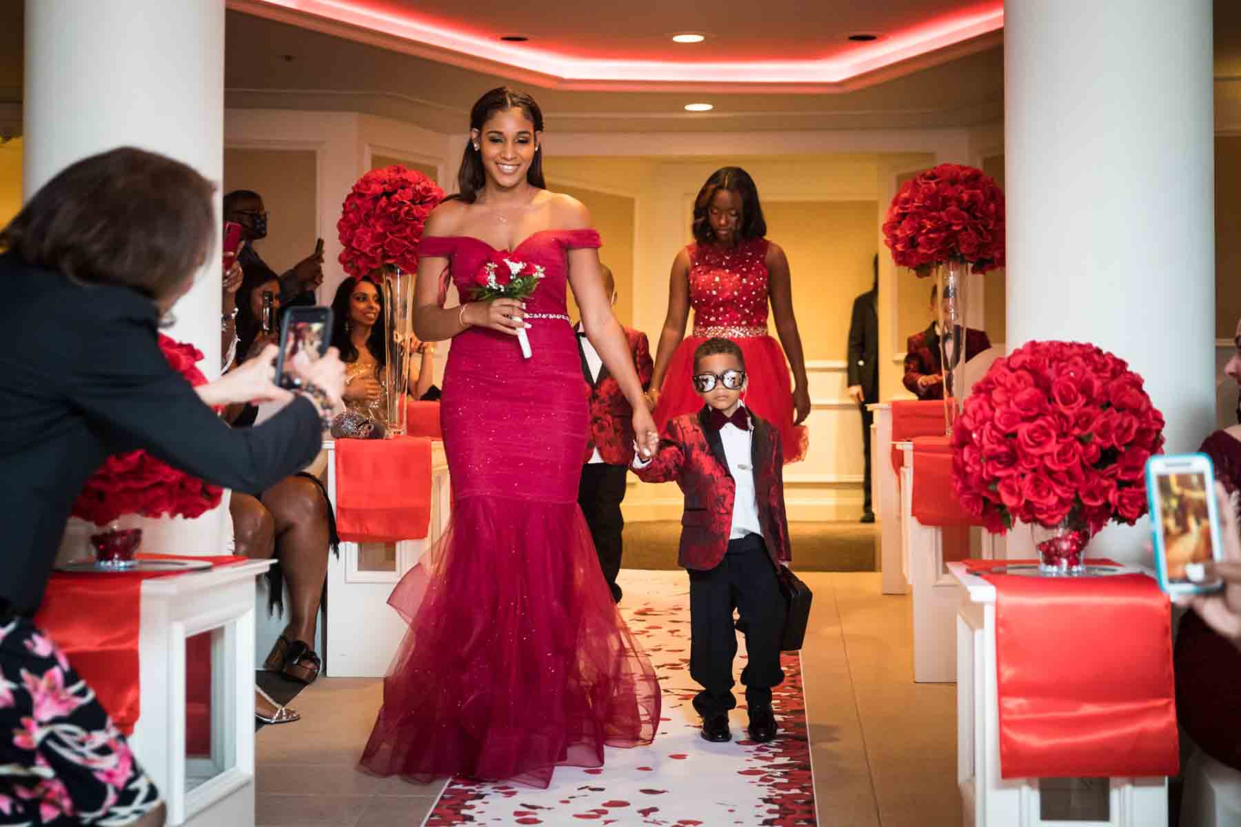 Woman in floor-length red dress leading little ring bearer down aisle for an article on the best wedding jobs for family and friends