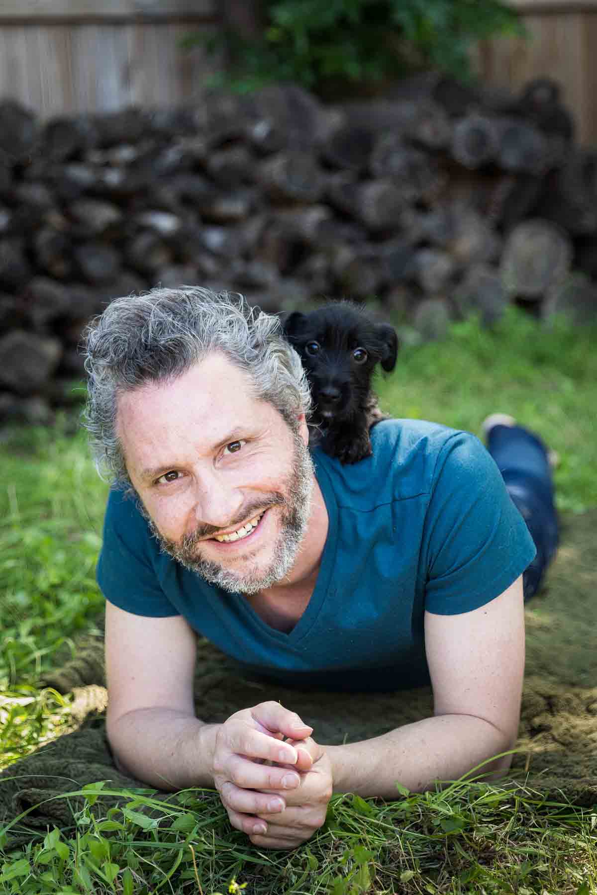 Man with gray hair, beard, and blue shirt lying on ground with black puppy on his shoulder for an article on pet photo tips