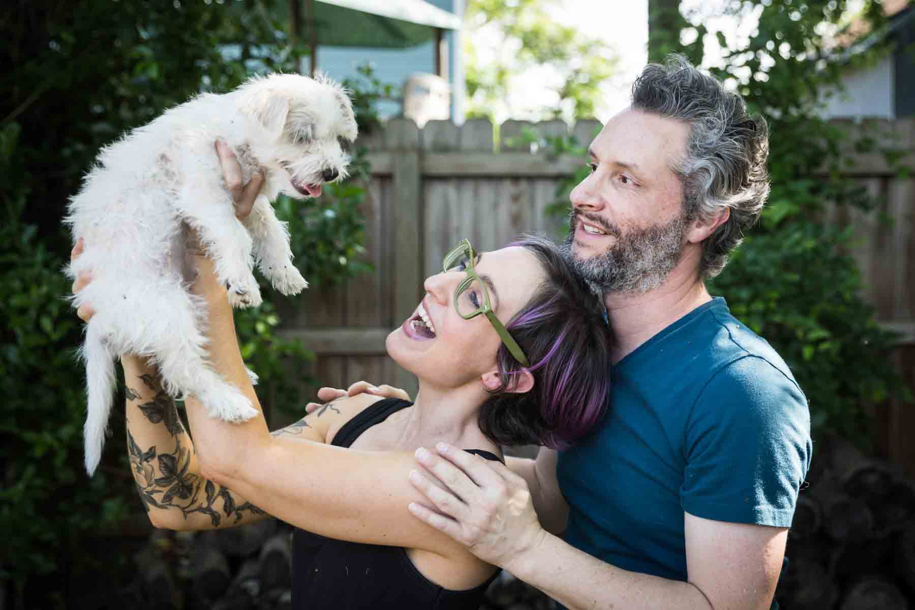 Woman holding up white puppy in front of man and wooden fence for an article on pet photo tips