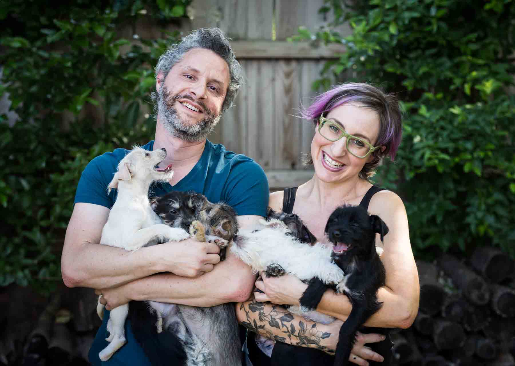 Man and woman holding puppies in their arms in front of wooden fence for an article on pet photo tips