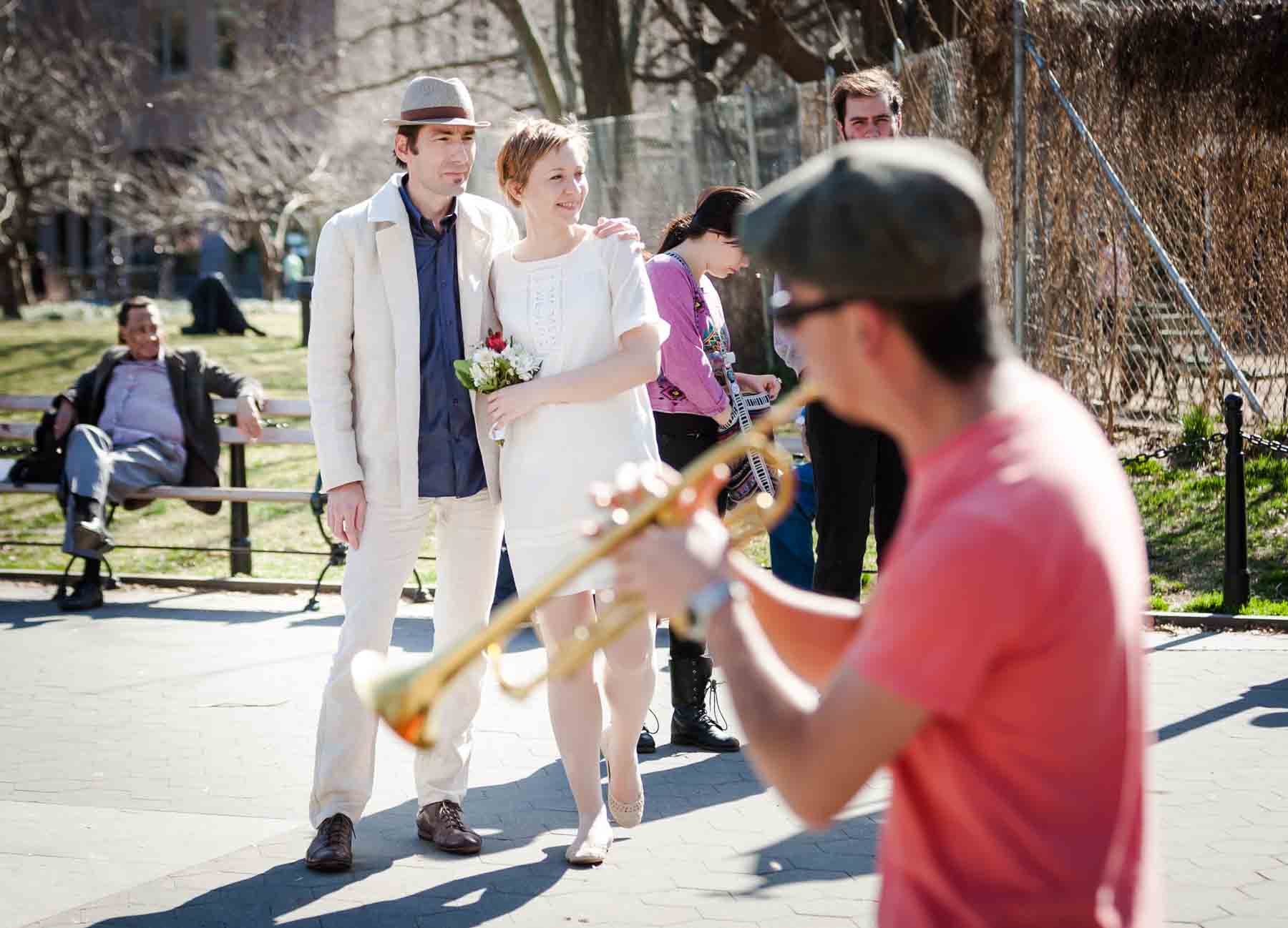 Couple standing close walking in park with man playing musical instrument in front for an article entitled, ‘Are Couples Still Getting Married?’