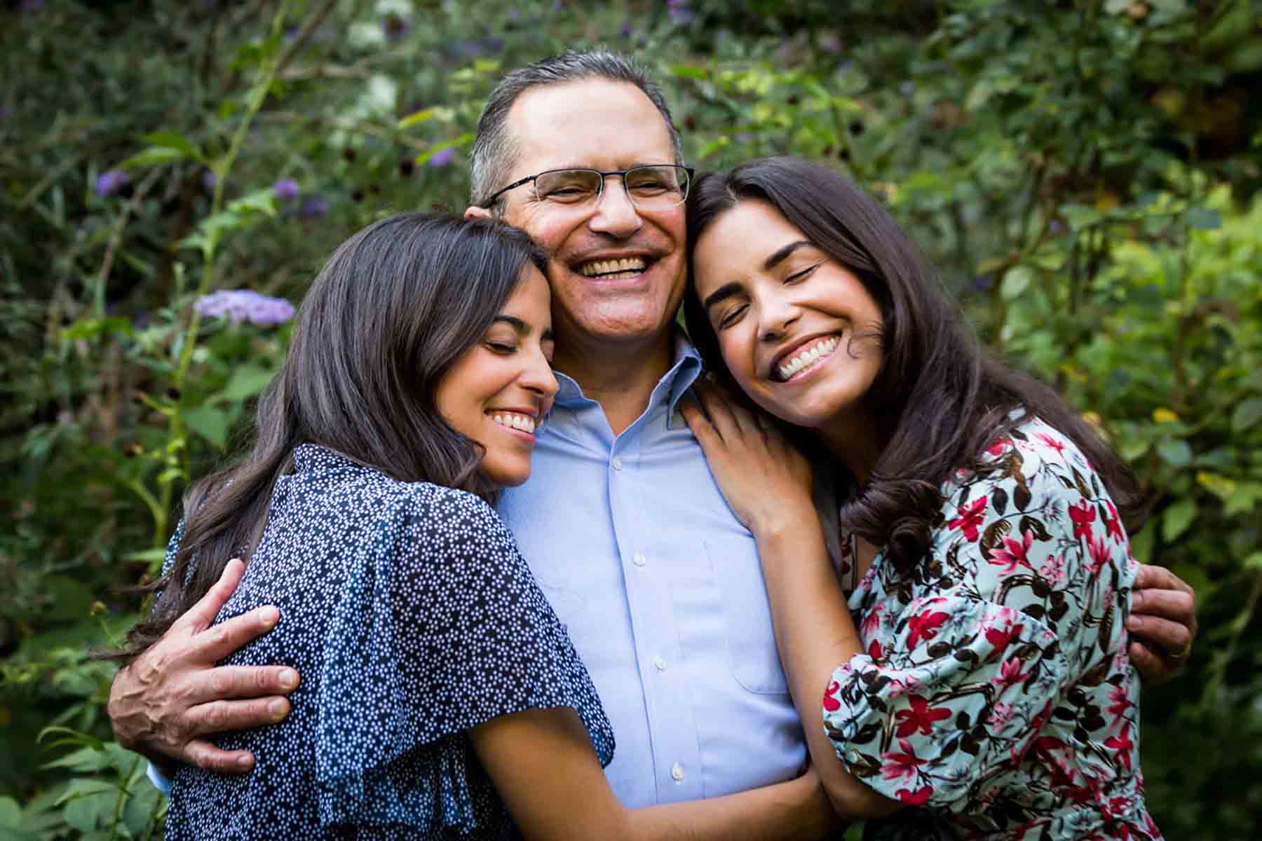 Two daughters hugging father in park in front of green bushes for an article on daddy and me photo shoot ideas