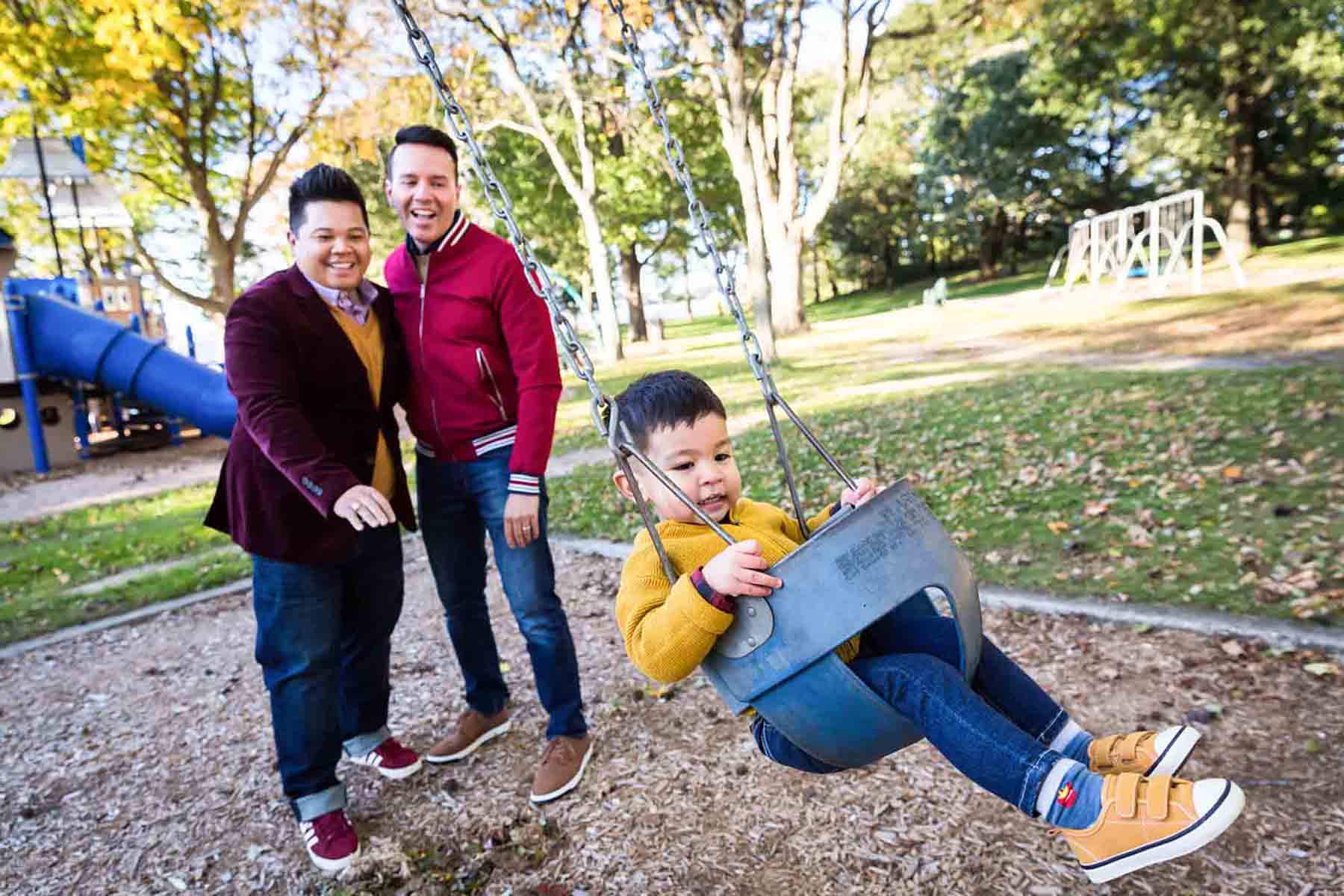 Two gay fathers pushing little boy in swing at playground for an article on daddy and me photo shoot ideas